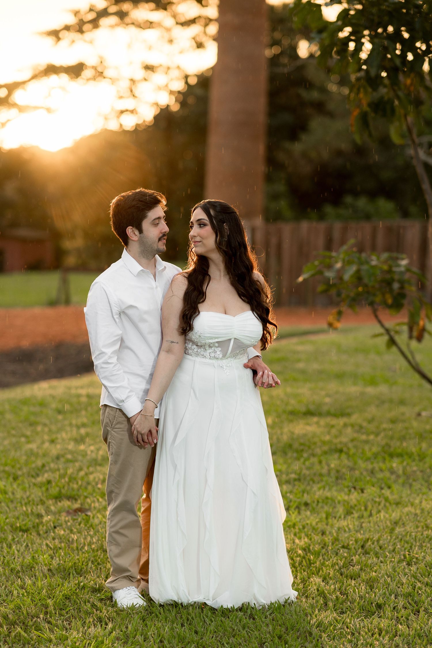Couple holding hands in white wedding attire at golden hour sunset in lush green garden.