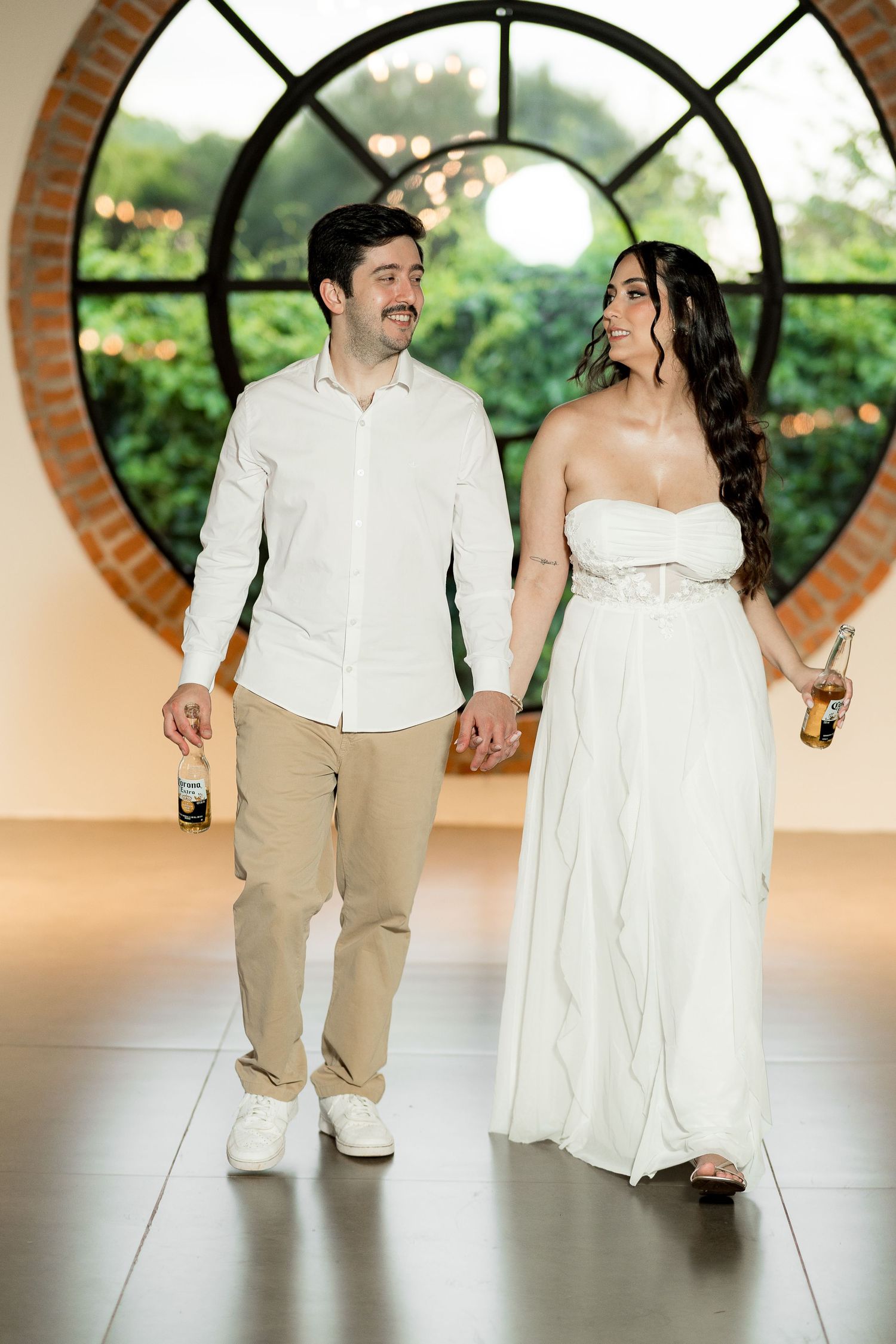 Couple in wedding attire holding hands and beer bottles walking in front of a circular brick window.