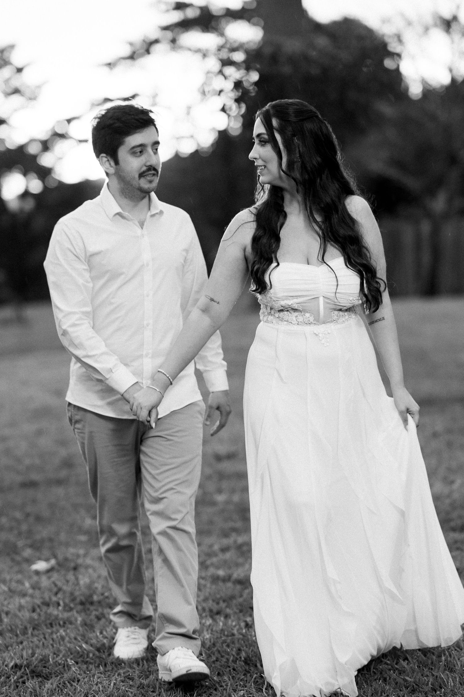 Black and white photo of a couple holding hands and walking in a park, woman in white gown smiling at man.