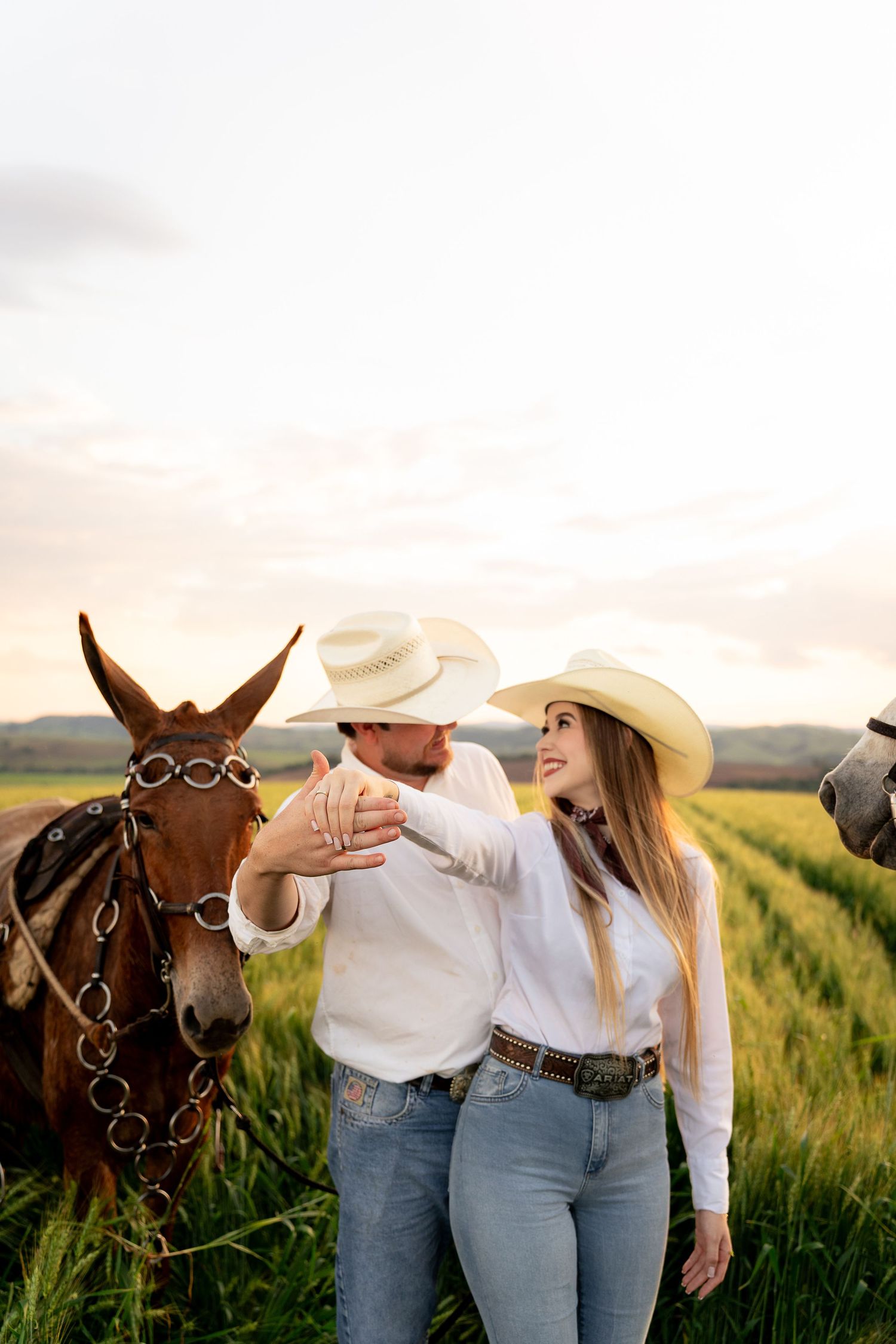 A couple in cowboy hats and jeans stand with a brown horse in a green field at sunset.