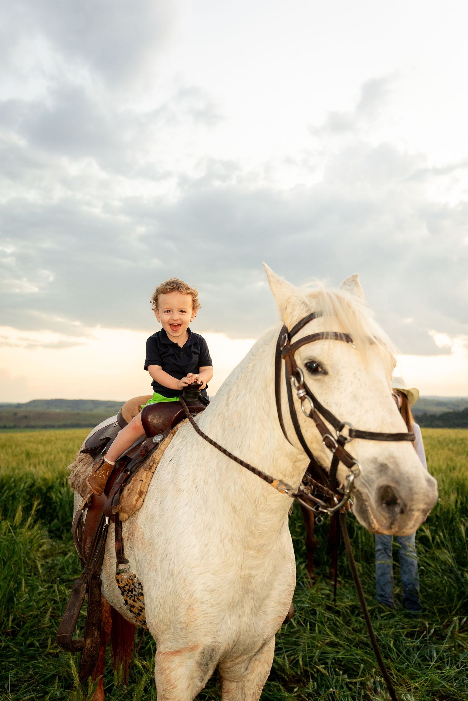 Young child smiling while riding a white horse in a green field under a cloudy sky at sunset.