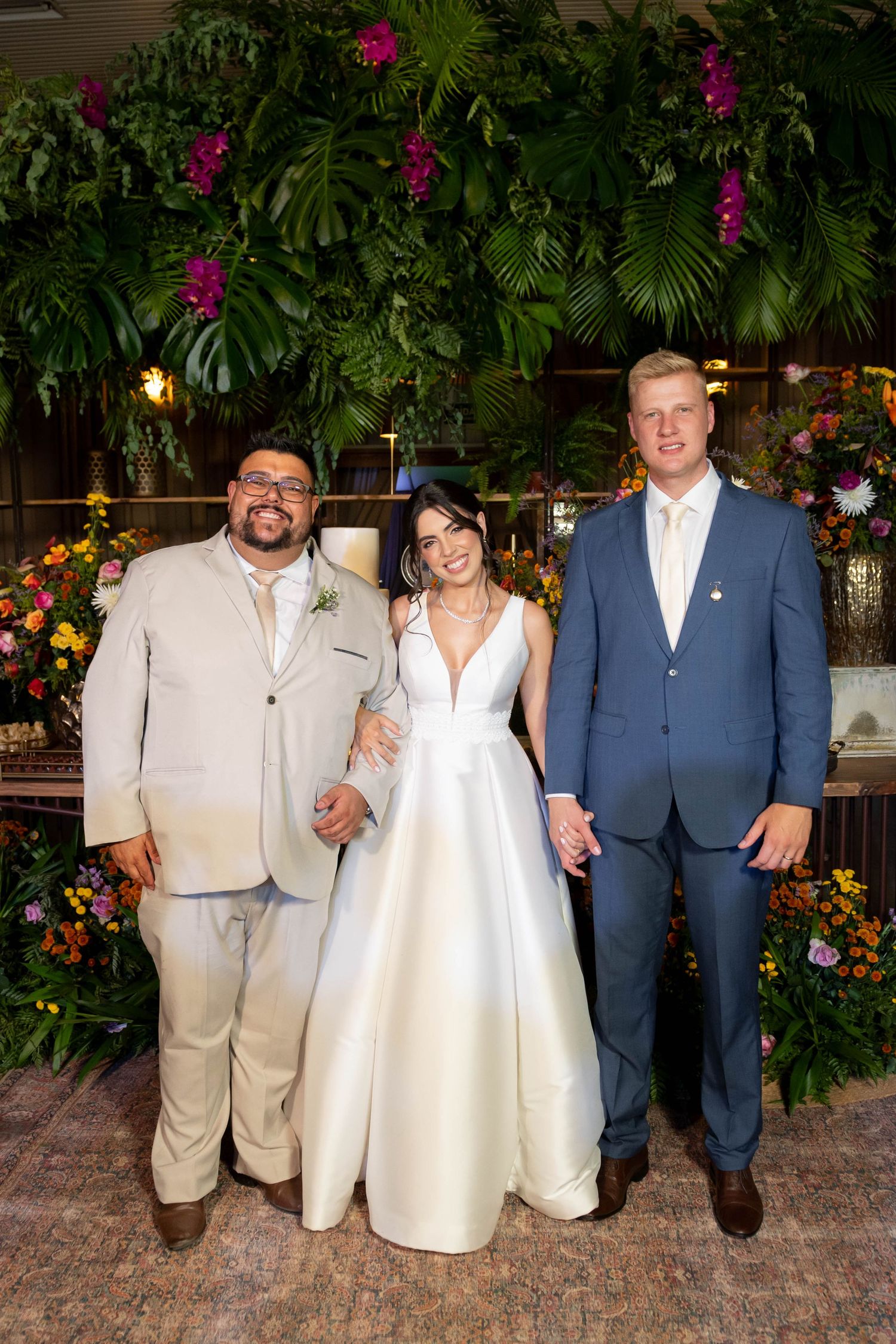Bride in white gown poses with two men in suits before a lush tropical floral backdrop at a wedding reception.