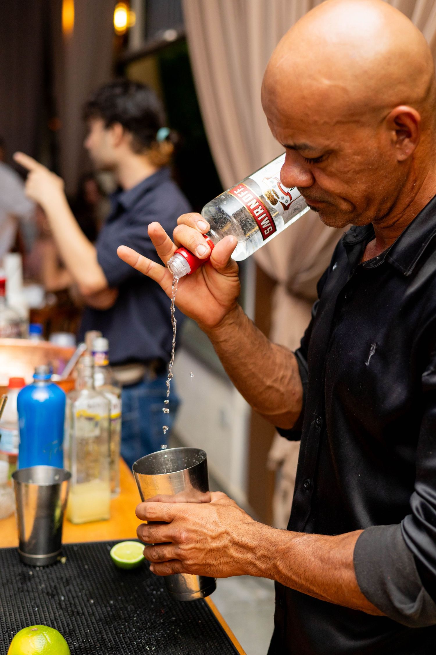 Bald bartender pouring Smirnoff vodka into a metal shaker at a bar event.