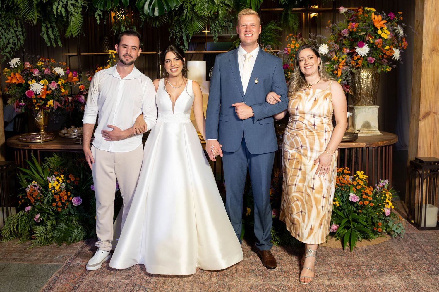 A bride in white gown poses with groom in blue suit and two guests amid colorful floral backdrop at wedding reception.