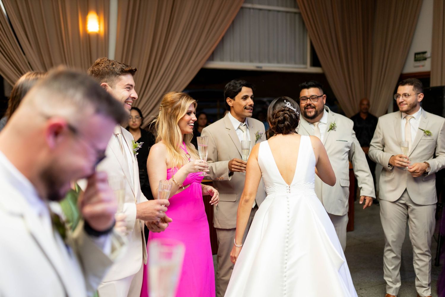 Wedding reception guests in beige suits and pink dress mingle with bride in white backless gown indoors.