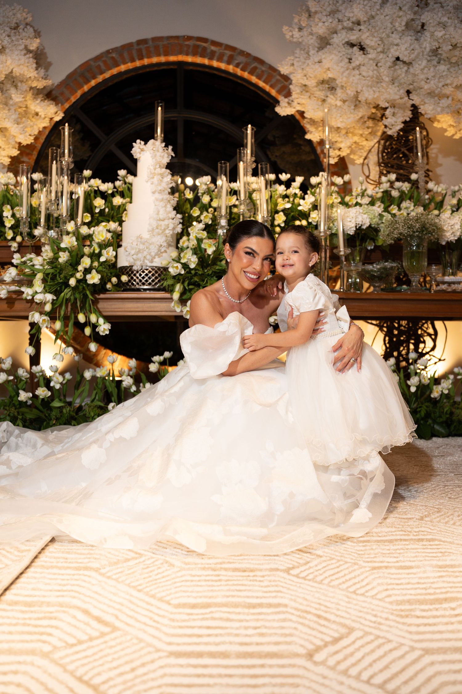 Bride in white ball gown poses with young flower girl in front of elegant floral wedding backdrop with tiered cake.