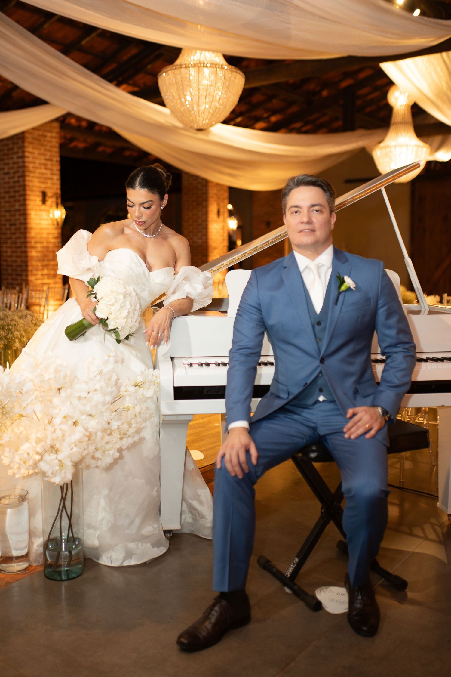 Bride in white gown with bouquet and groom in blue suit pose elegantly by a white piano at their wedding reception.