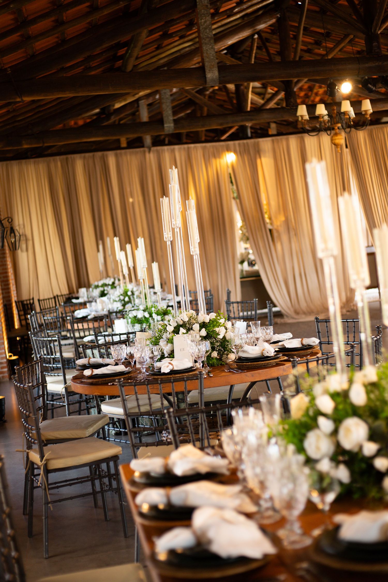 Elegant wedding reception venue with round tables, tall candelabras, white floral centerpieces, and draped fabric walls.