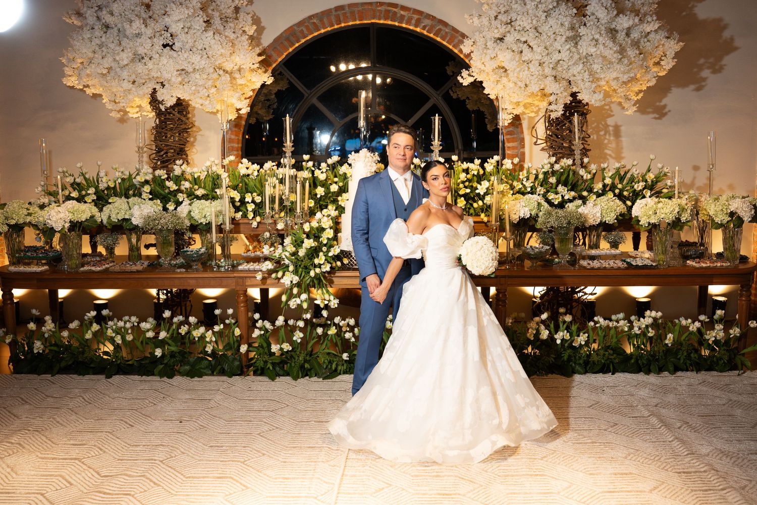 Bride and groom pose together in an elegant wedding venue adorned with lush white floral arrangements and candlelight.