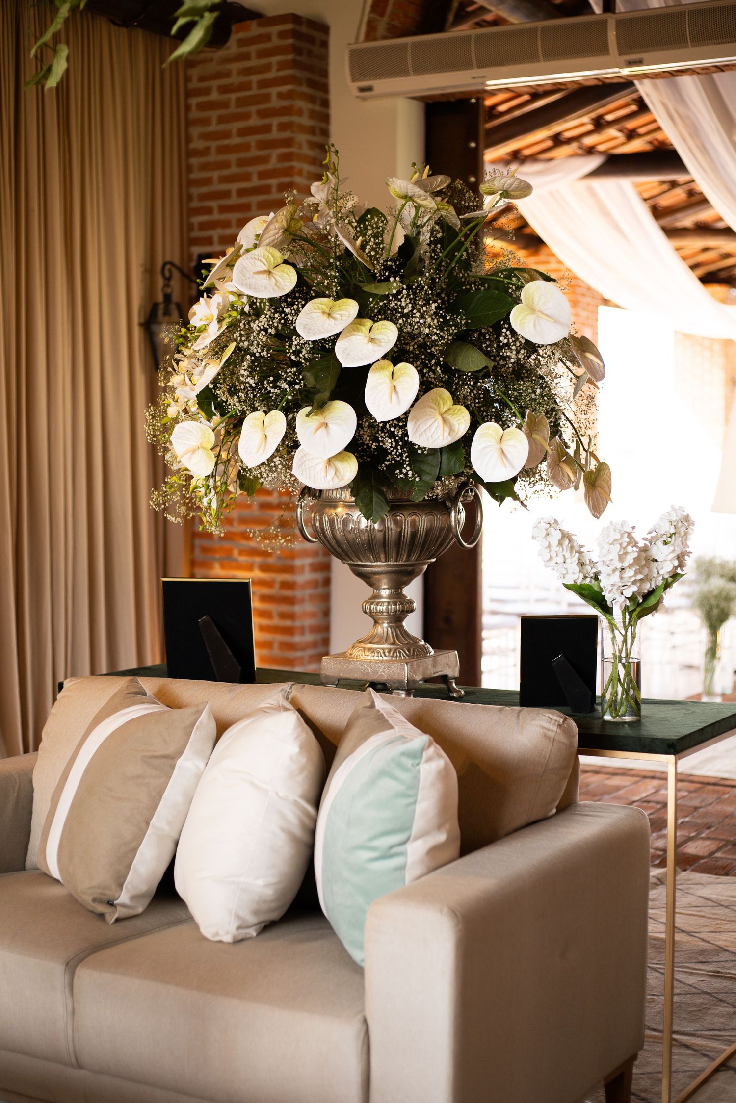Elegant event lounge with beige sofa, decorative pillows, and large floral arrangement of white anthuriums in silver urn.