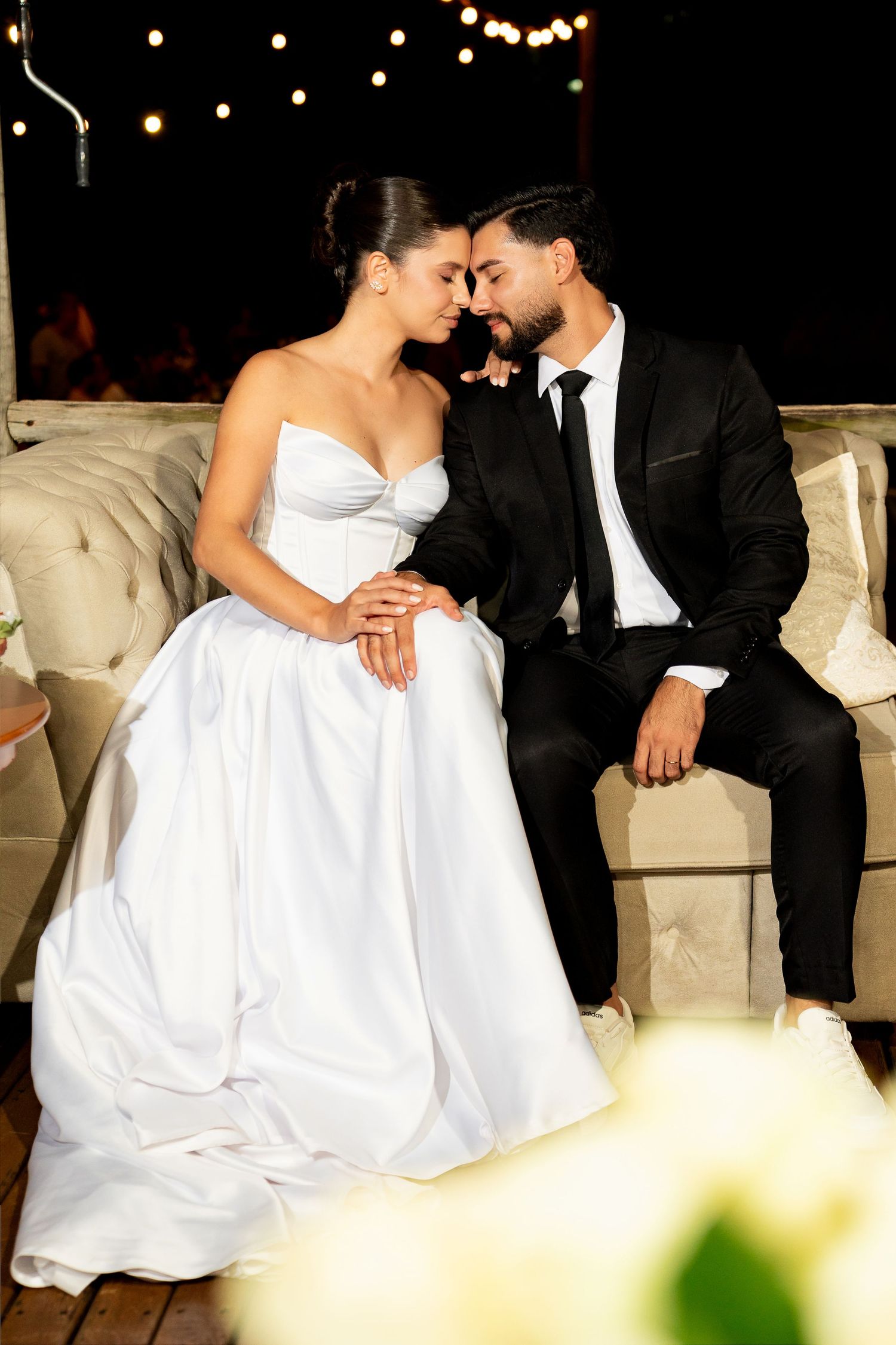 Bride in white strapless gown and groom in black suit sit closely together, foreheads touching at an outdoor evening wedding.