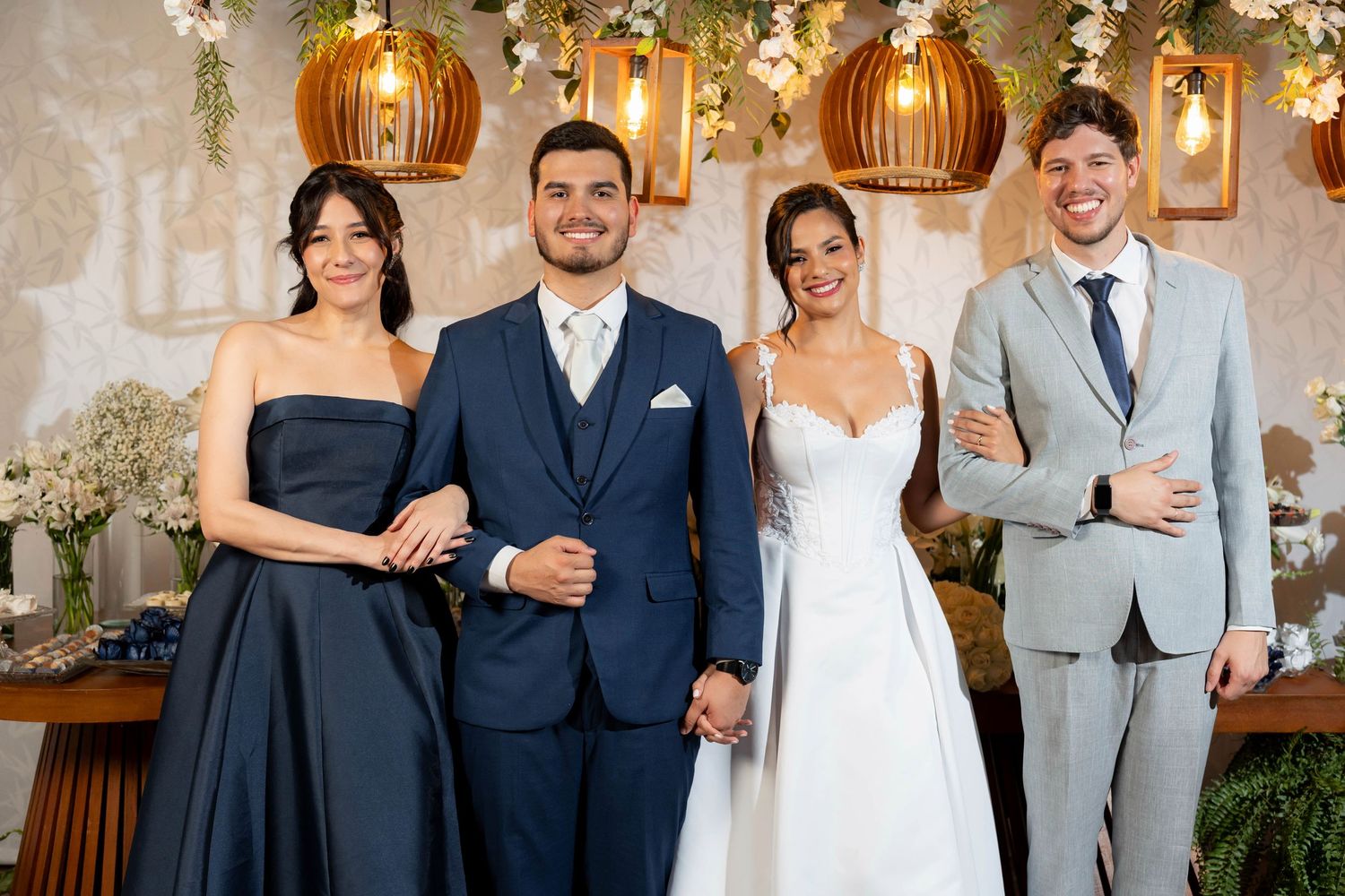 A bride and groom pose with two guests at a wedding reception, surrounded by floral decor and warm pendant lights.