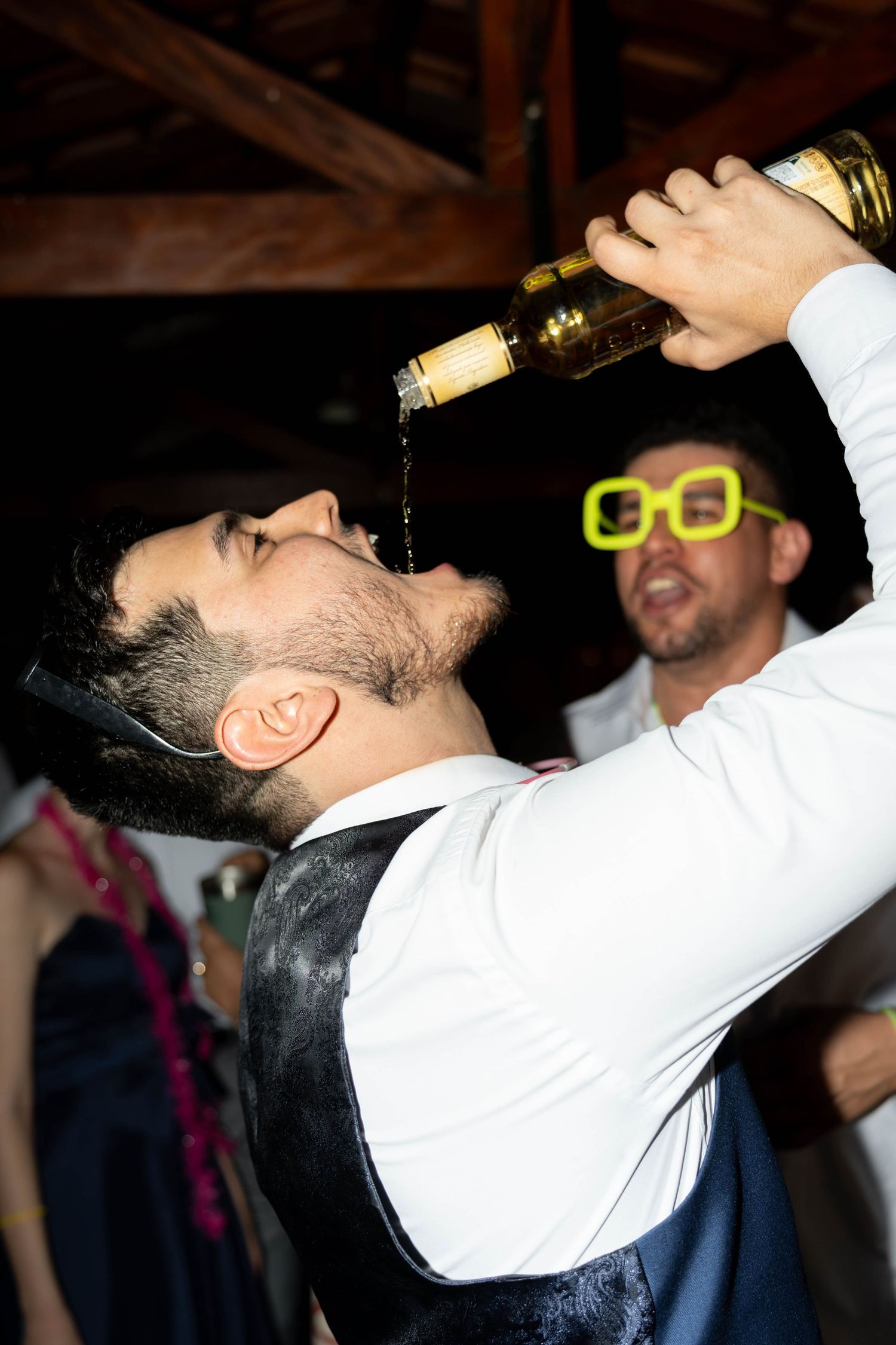Man in white shirt and vest pouring alcohol into his mouth at a party, with guest wearing neon glasses behind him.