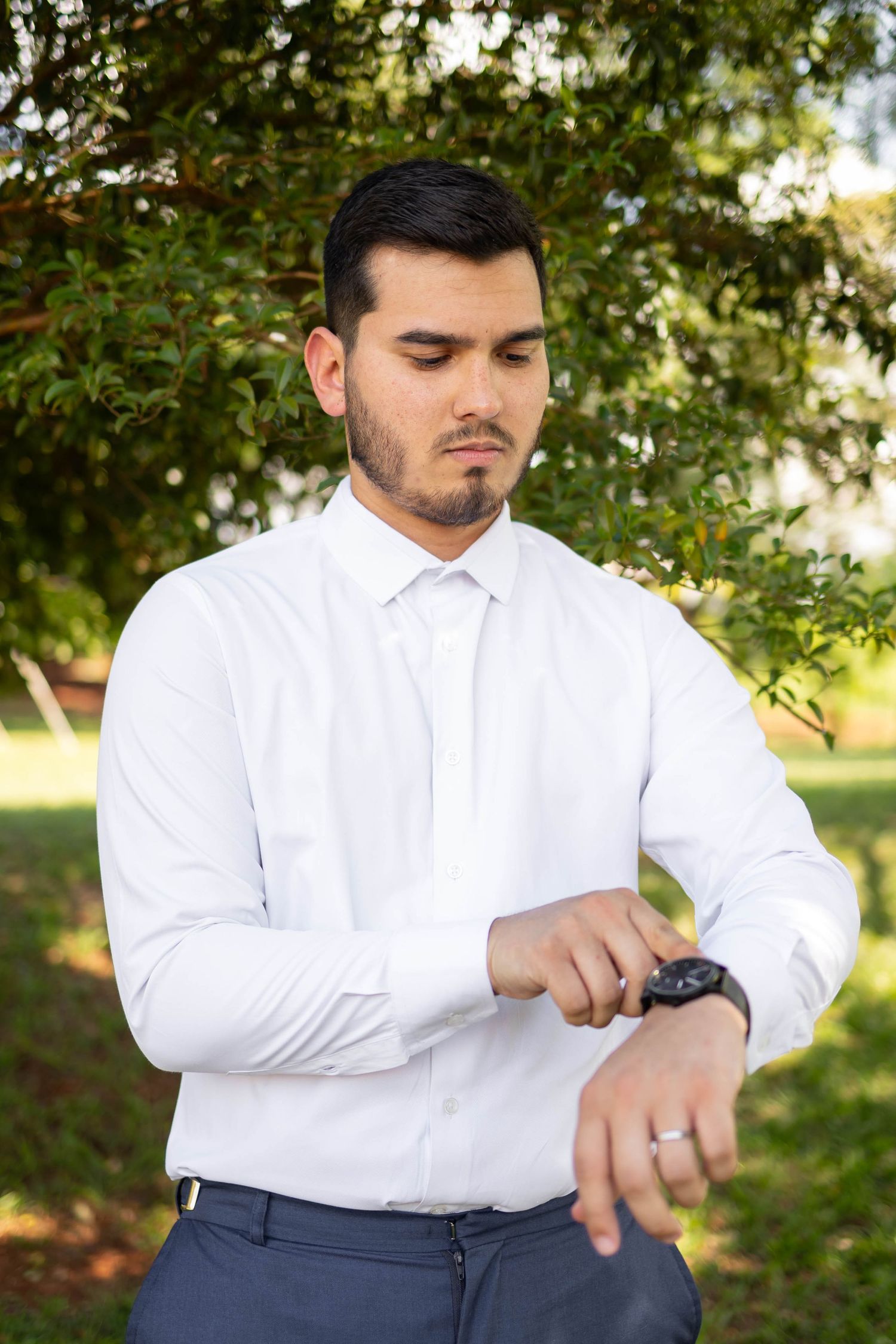 Young businessman in white shirt checking his wristwatch outdoors with green trees in background.