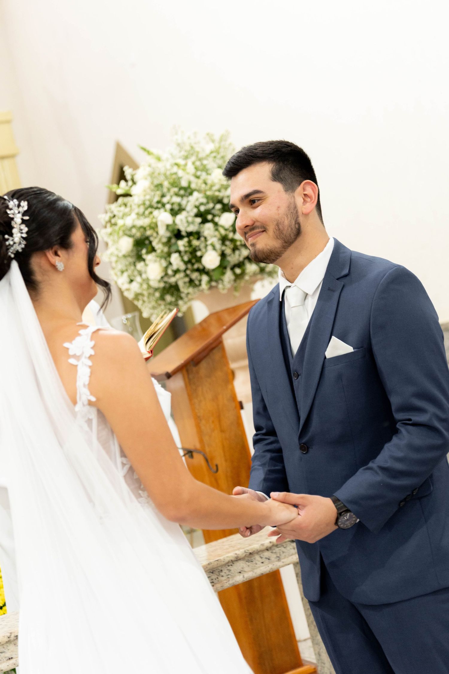 Bride and groom holding hands at the altar during a church wedding ceremony, with white floral arrangements in the background.