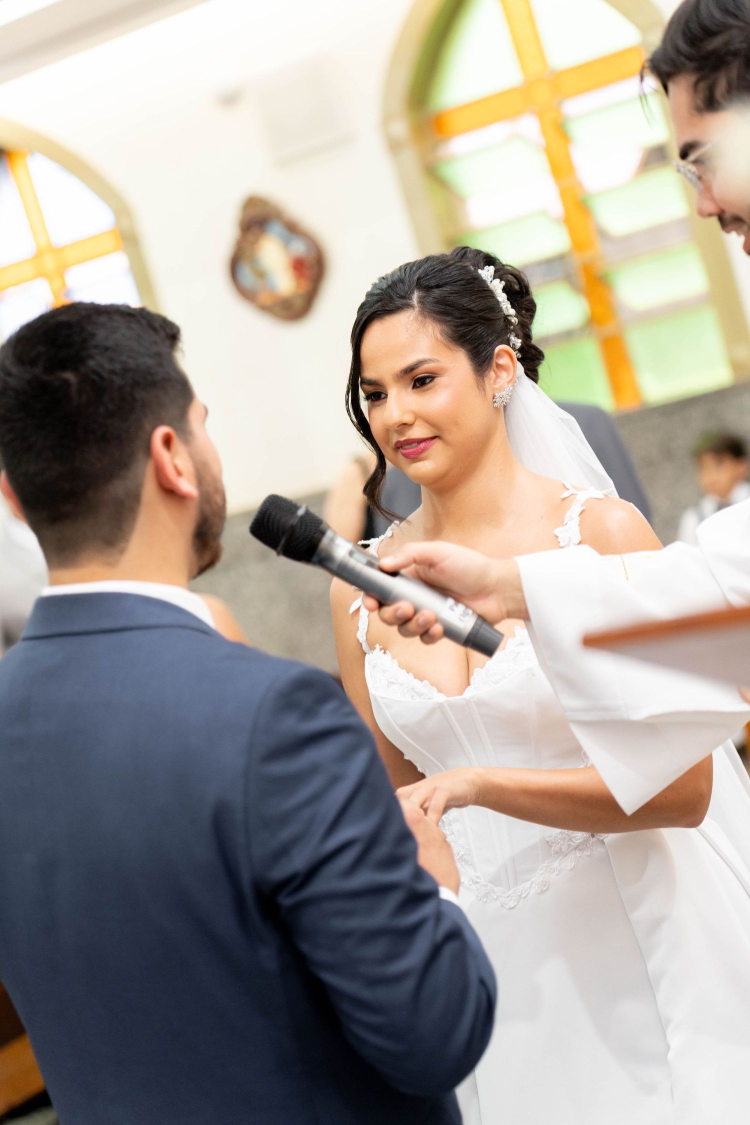Bride reciting wedding vows at church ceremony while officiant holds microphone, groom standing opposite in blue suit.