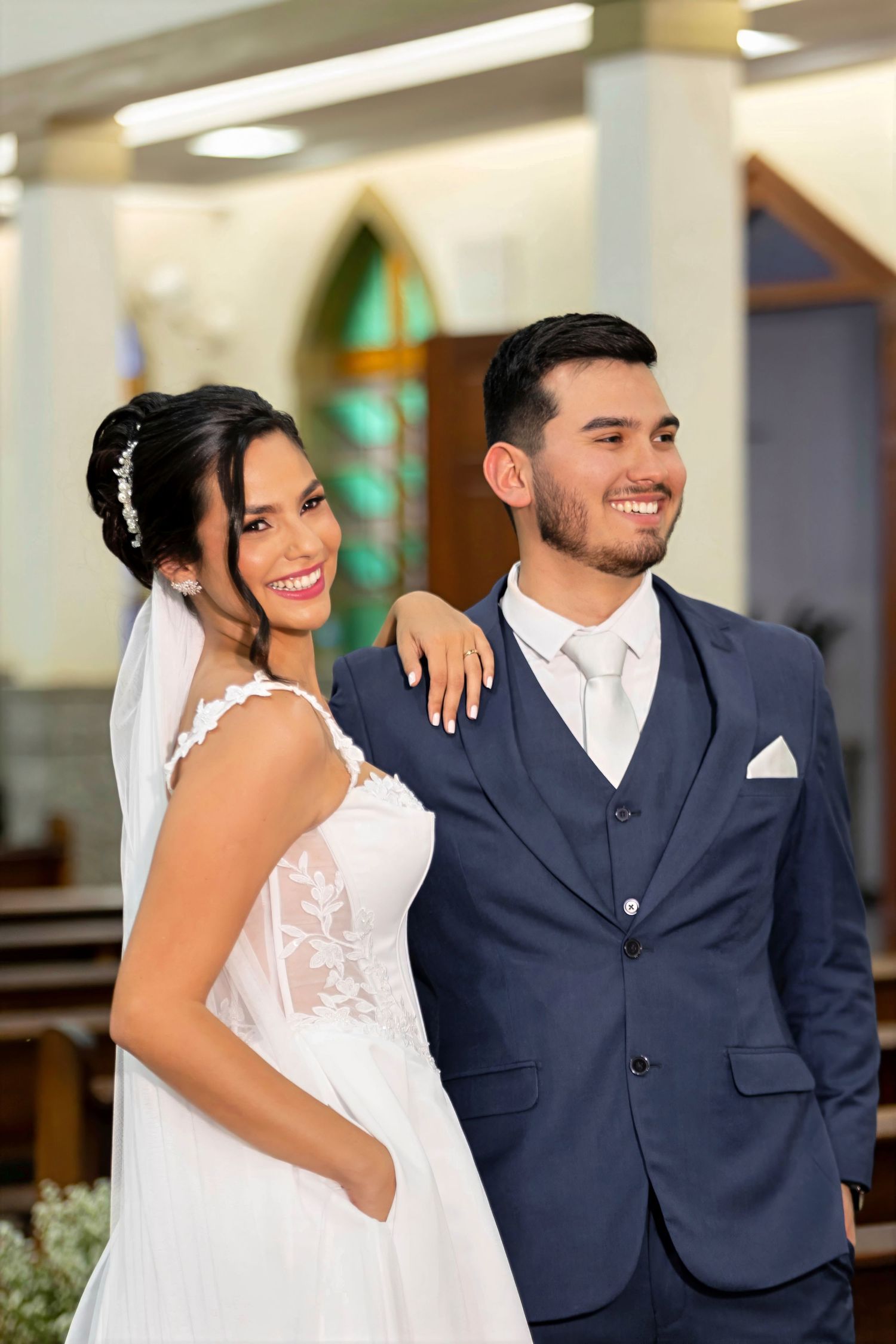 Smiling bride in white lace gown and veil stands beside groom in navy blue suit inside a church.