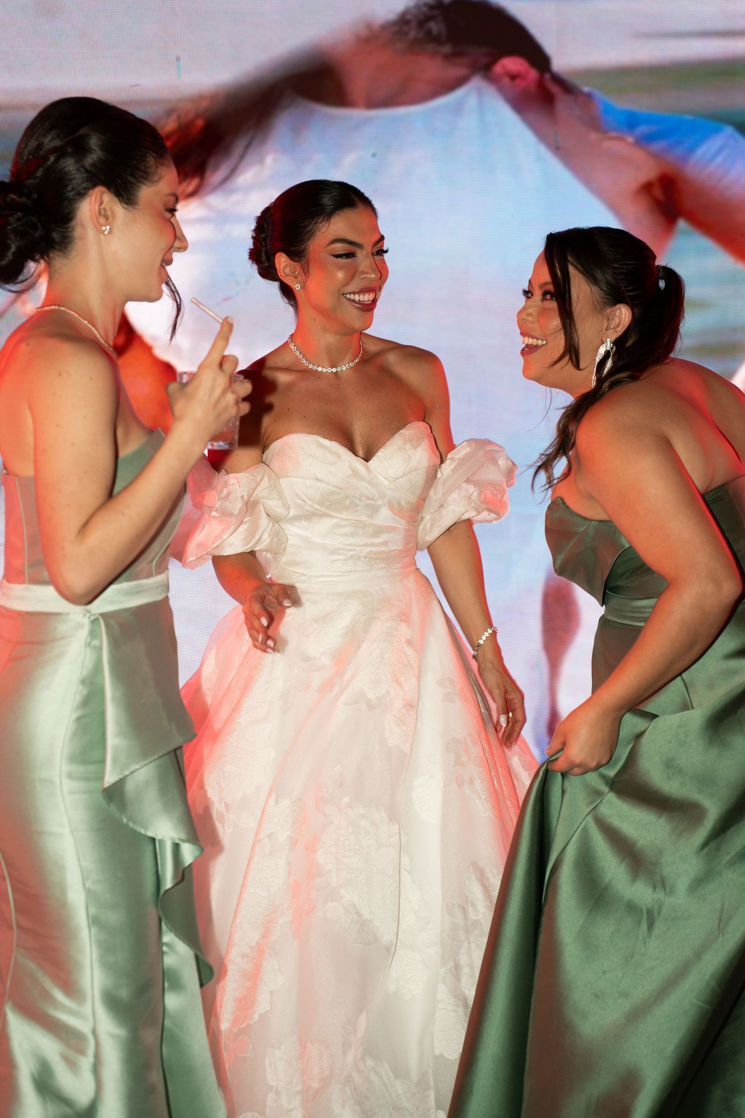 Bride in white gown laughs with bridesmaids in sage green satin dresses at a wedding reception.