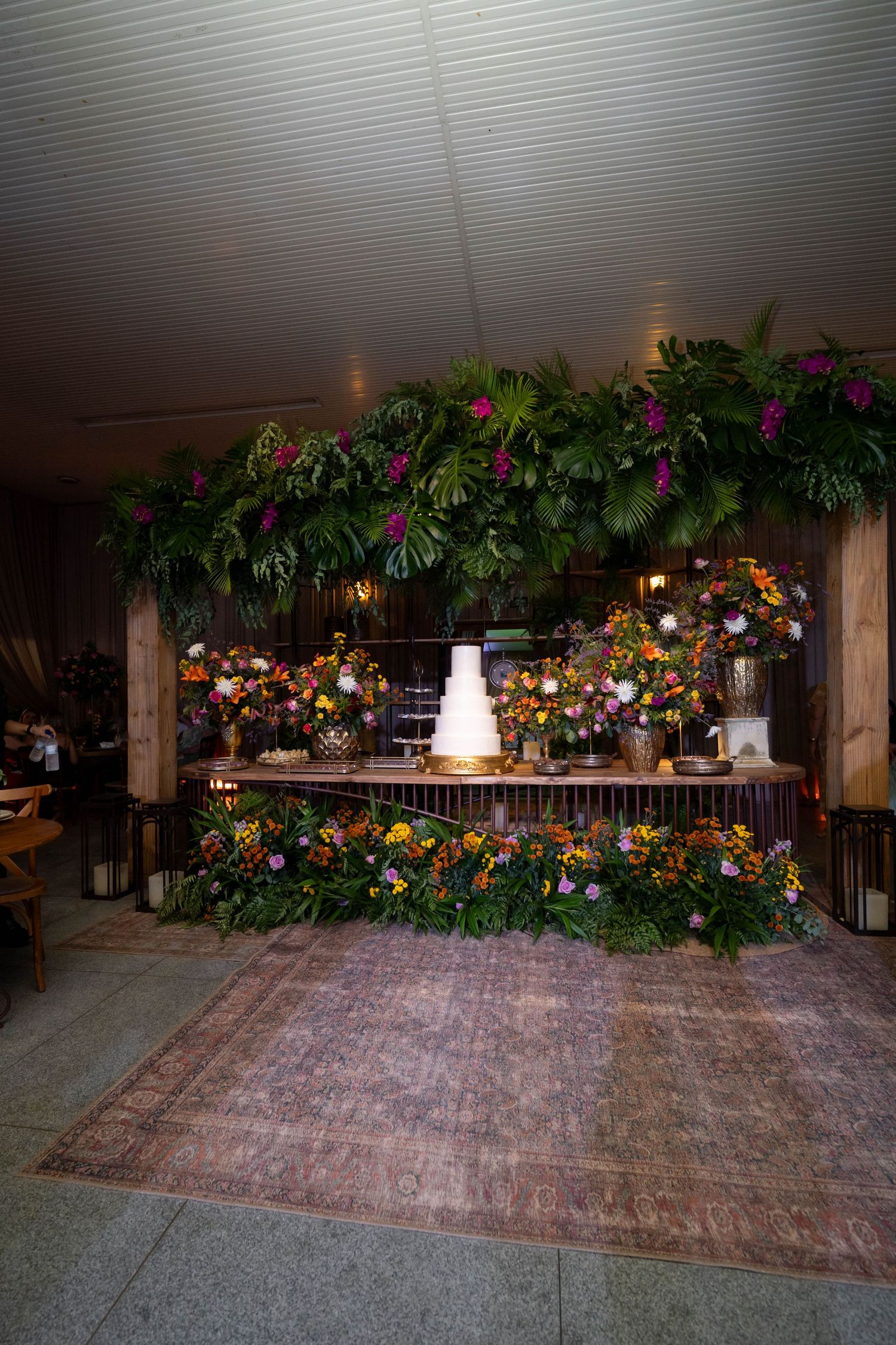 Elegant wedding cake display surrounded by lush tropical greenery and colorful floral arrangements on a rustic decorated table.