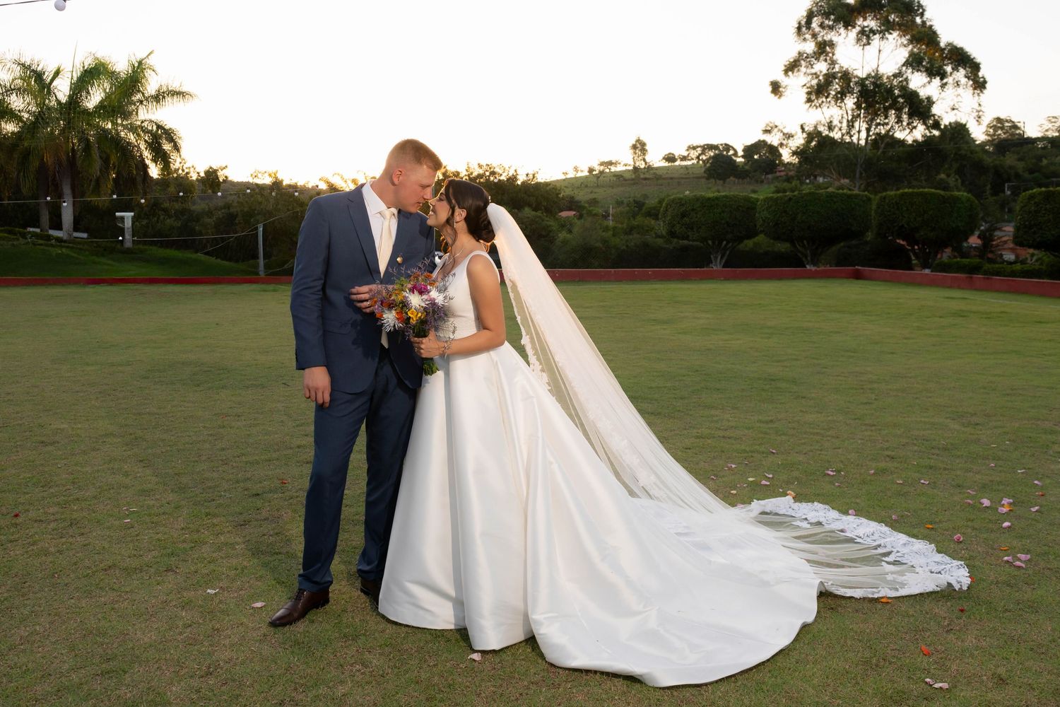 Bride and groom share a romantic moment on a lush green golf course at sunset, bride wearing a flowing white gown with veil.