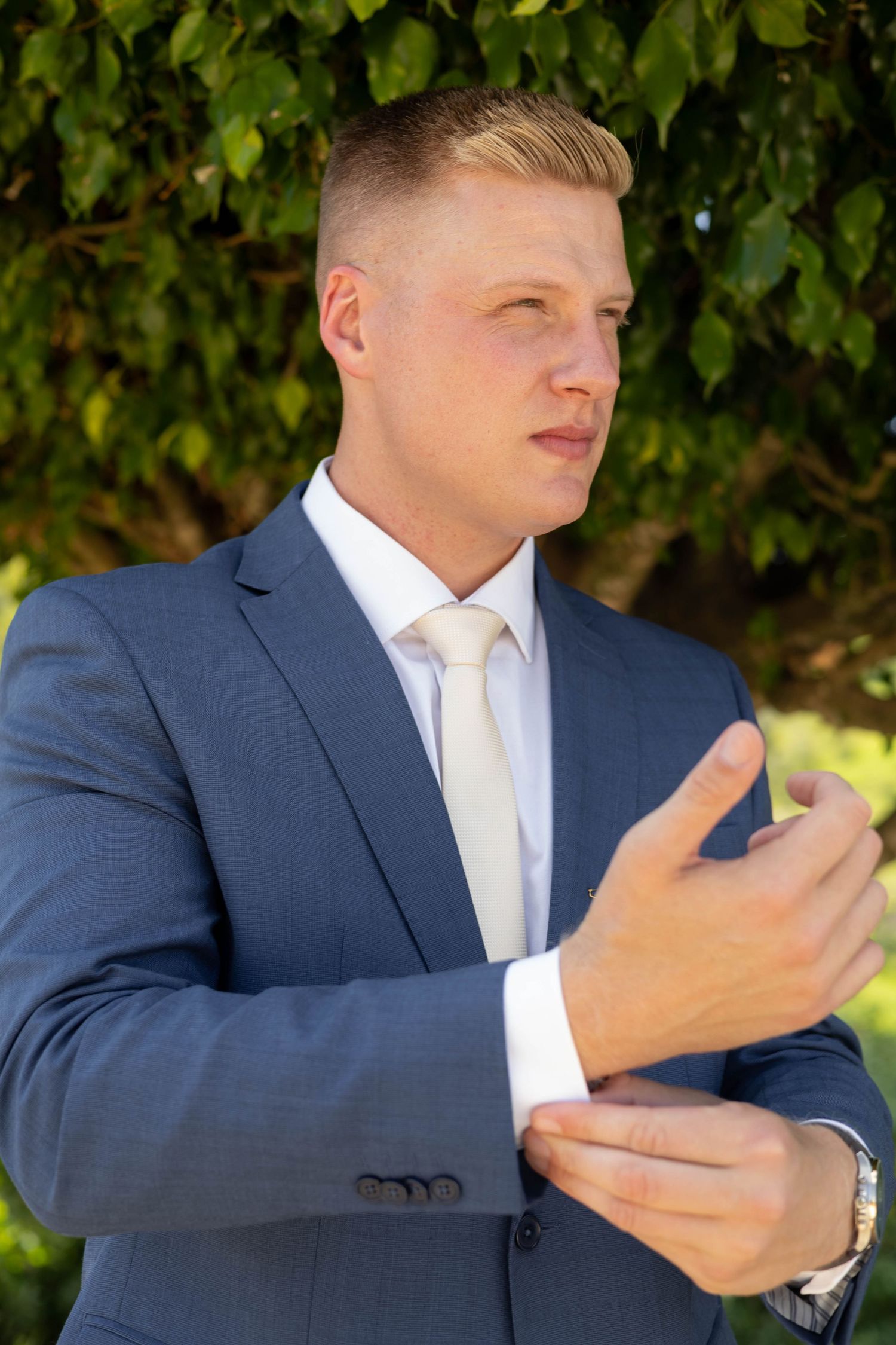 Young blond man in blue suit adjusting cufflinks outdoors with green foliage background.