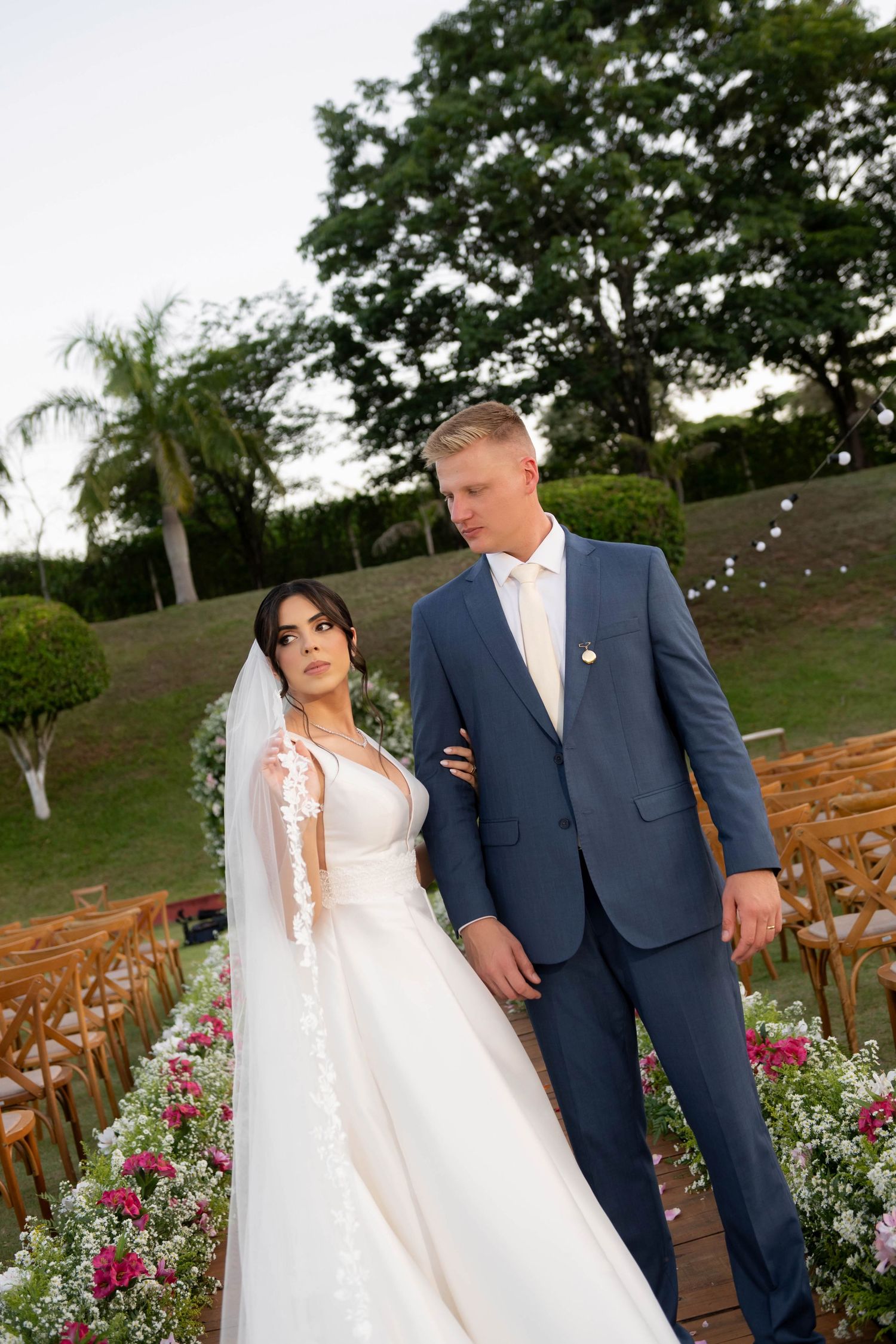 Bride in white gown and groom in blue suit stand together at outdoor wedding ceremony surrounded by floral arrangements.