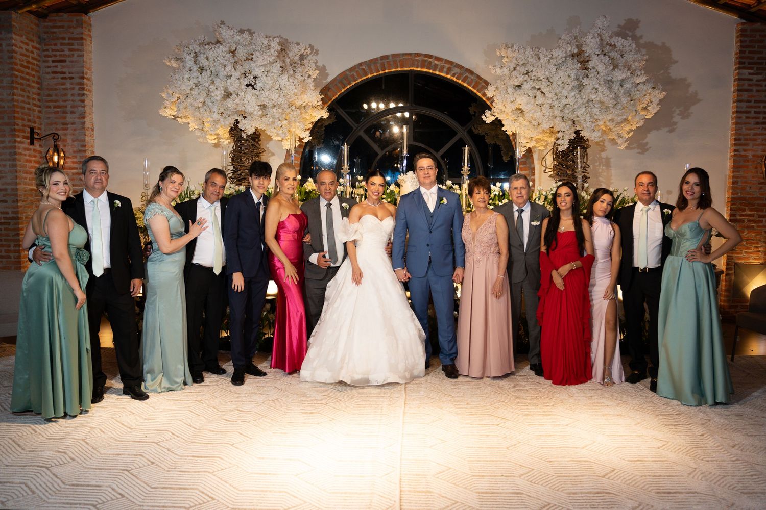 Wedding party poses together in elegant venue with floral arches, bride in white gown surrounded by guests in formal attire.