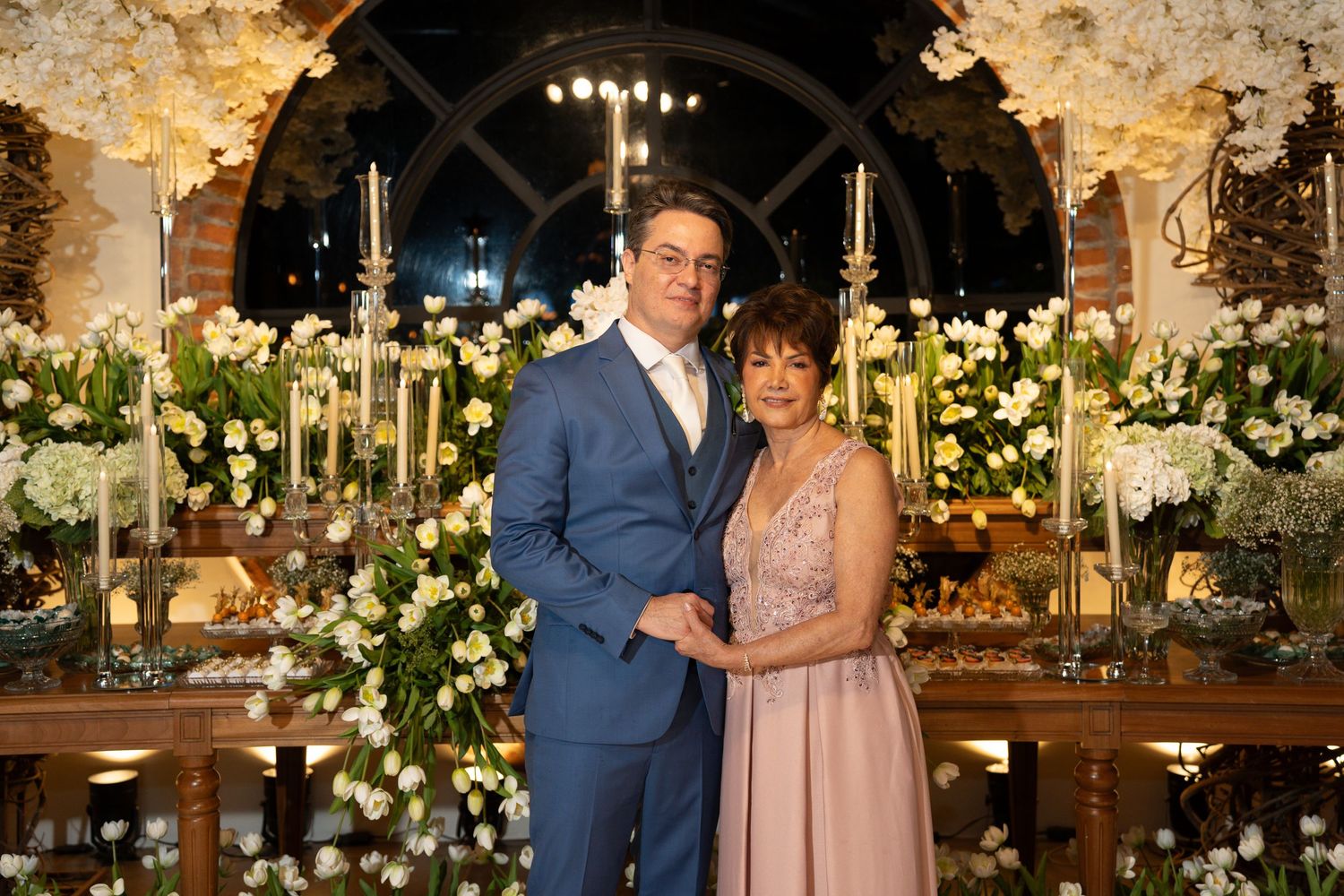 Elegant couple posing at a wedding reception with white floral arrangements and candlesticks in the background.