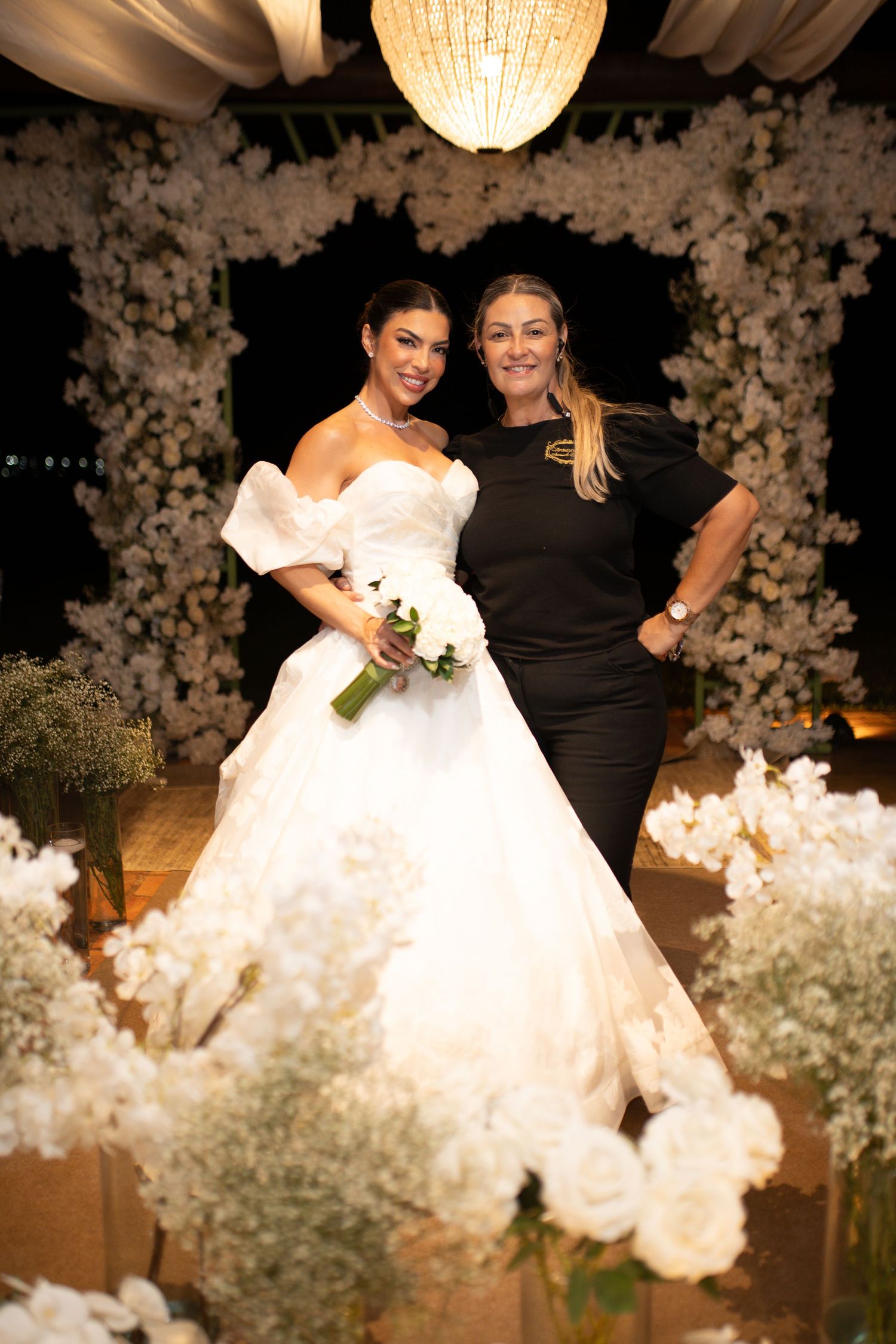 Bride in white ballgown holding bouquet poses with woman in black dress amid floral arch and white flower arrangements.