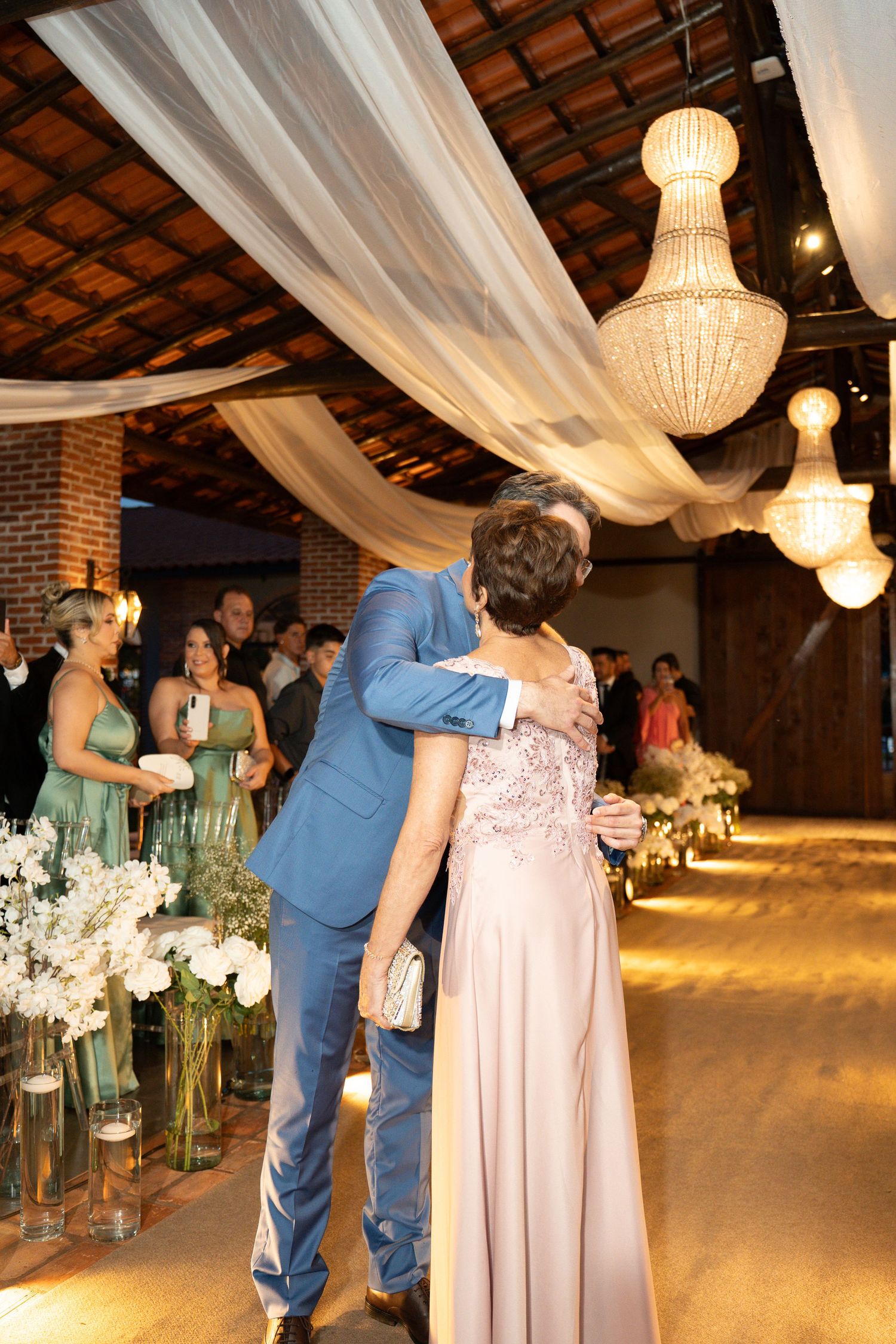 Groom in blue suit embraces woman in pink gown at elegant wedding venue with chandeliers and floral decor.