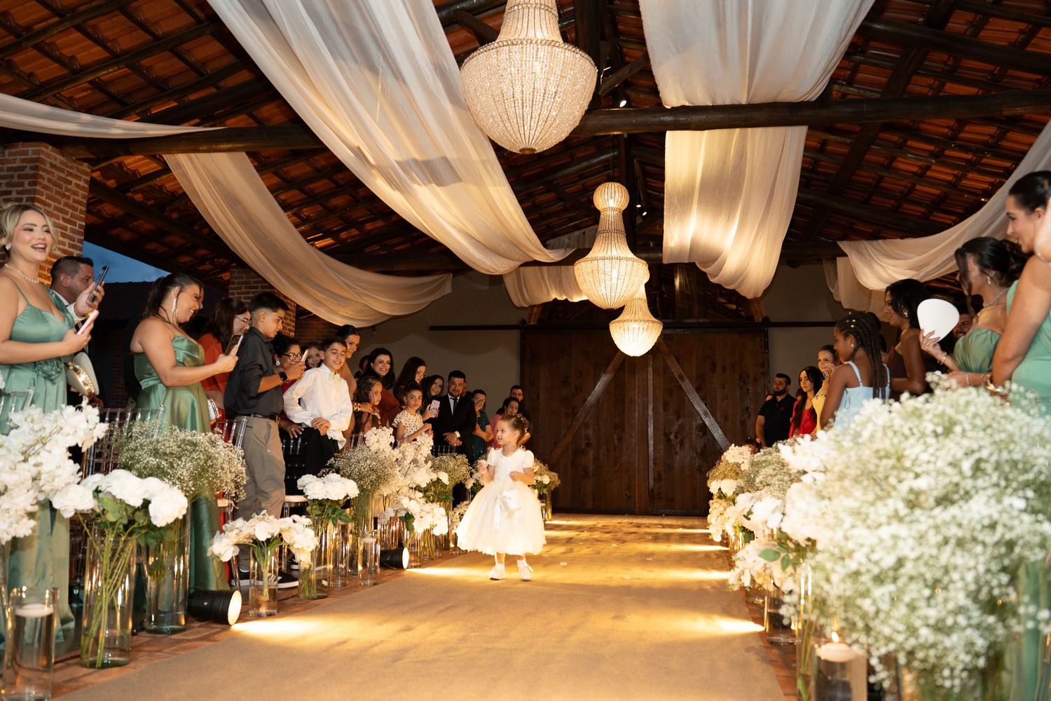 Flower girl walks down a floral-lined wedding aisle under draped white fabric ceiling with chandelier lighting.