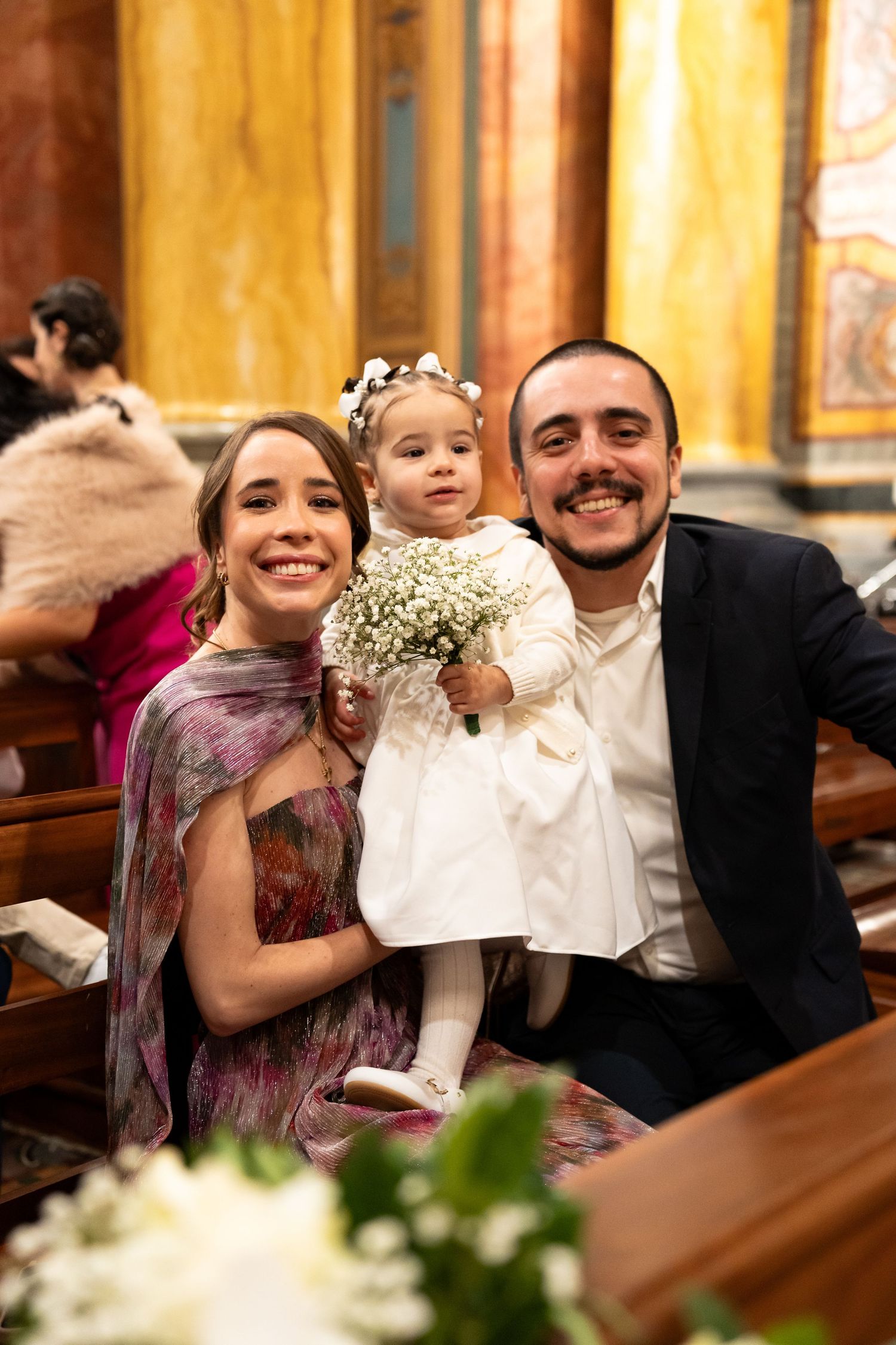 Family posing in church pew, smiling girl in white dress holding baby's breath bouquet, flanked by parents.