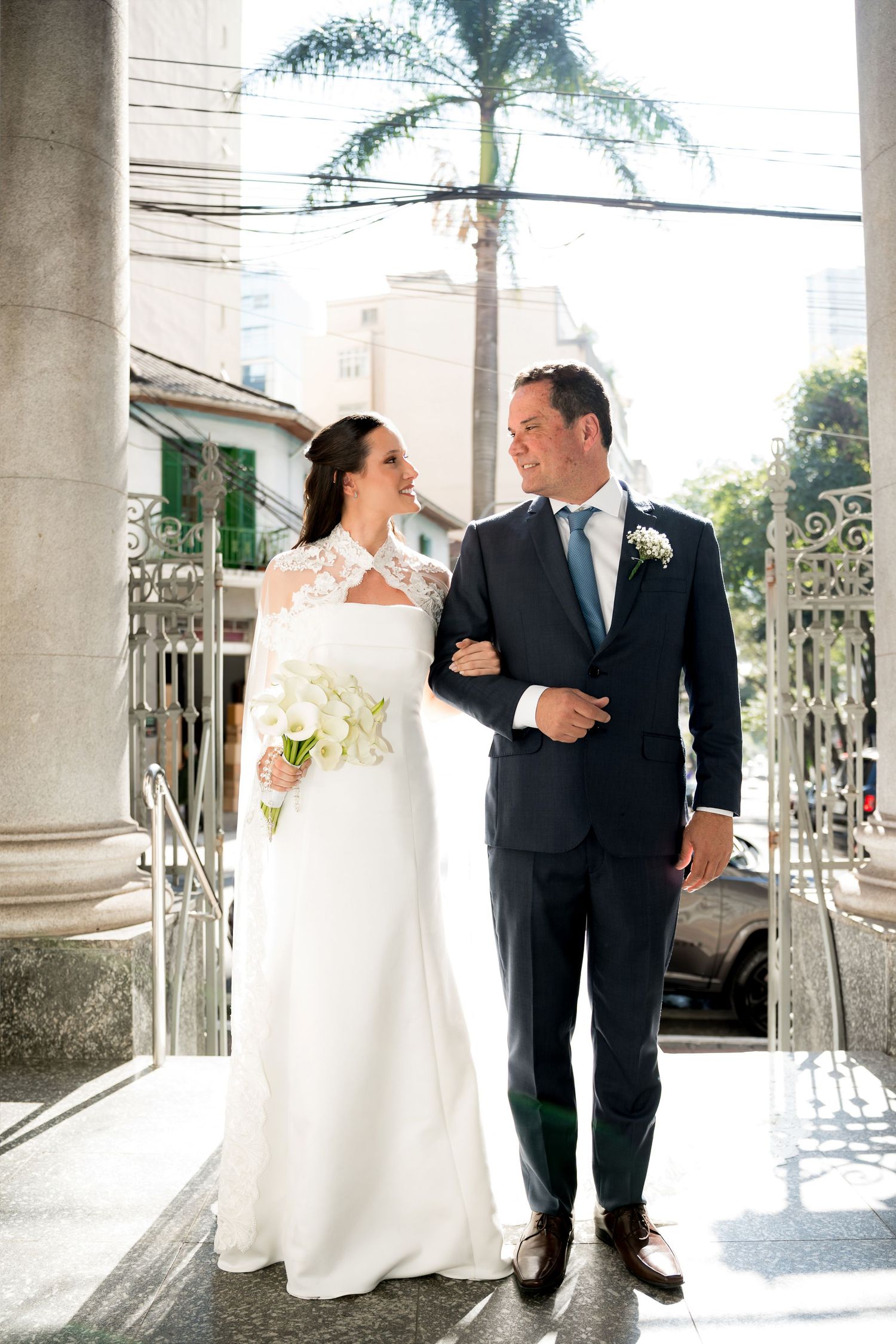 Bride in white lace gown holding bouquet walks arm-in-arm with groom in navy suit outside ornate church gates.