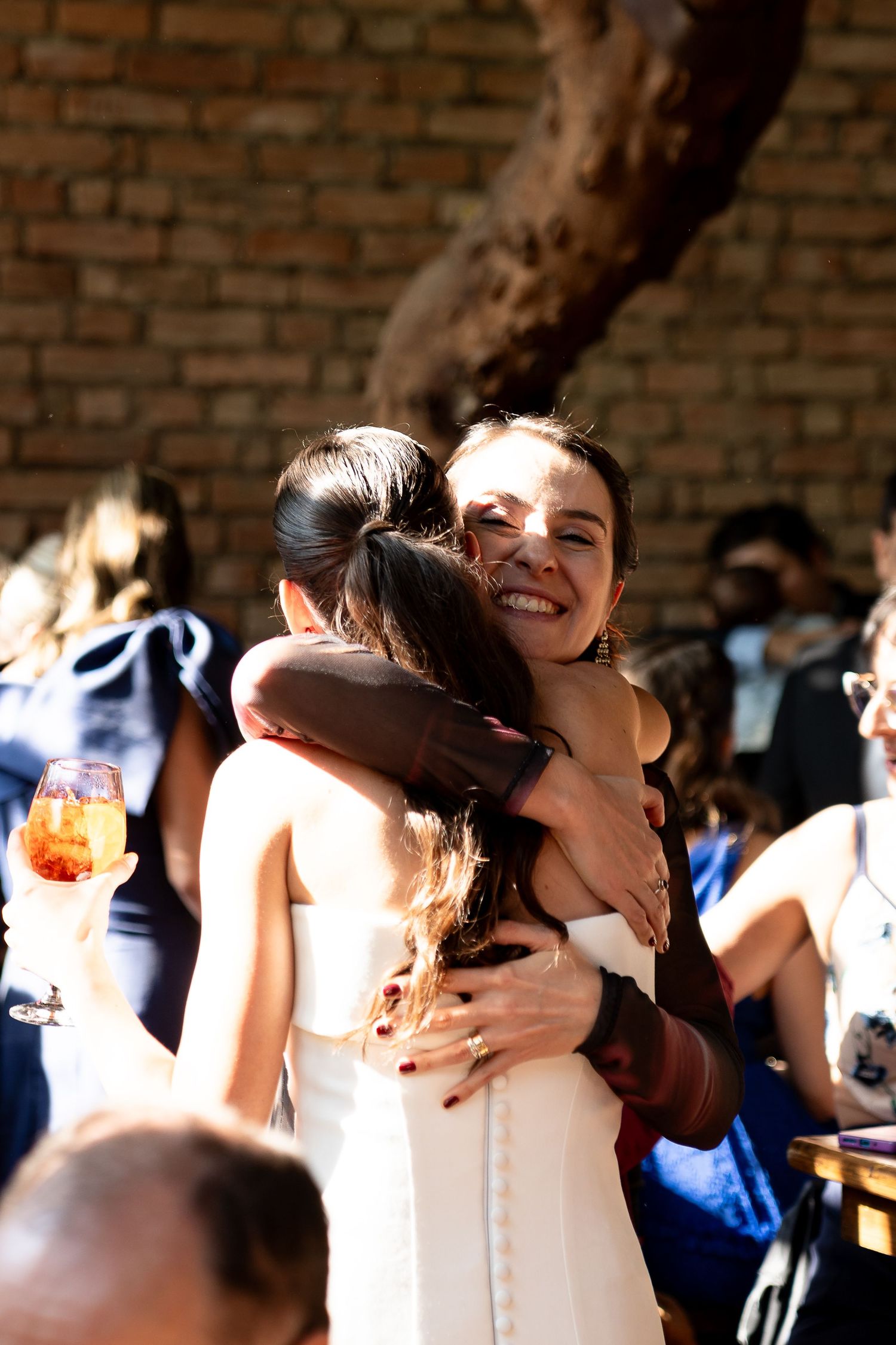 Two women sharing a warm hug at an outdoor wedding celebration, smiling joyfully with guests in the background.
