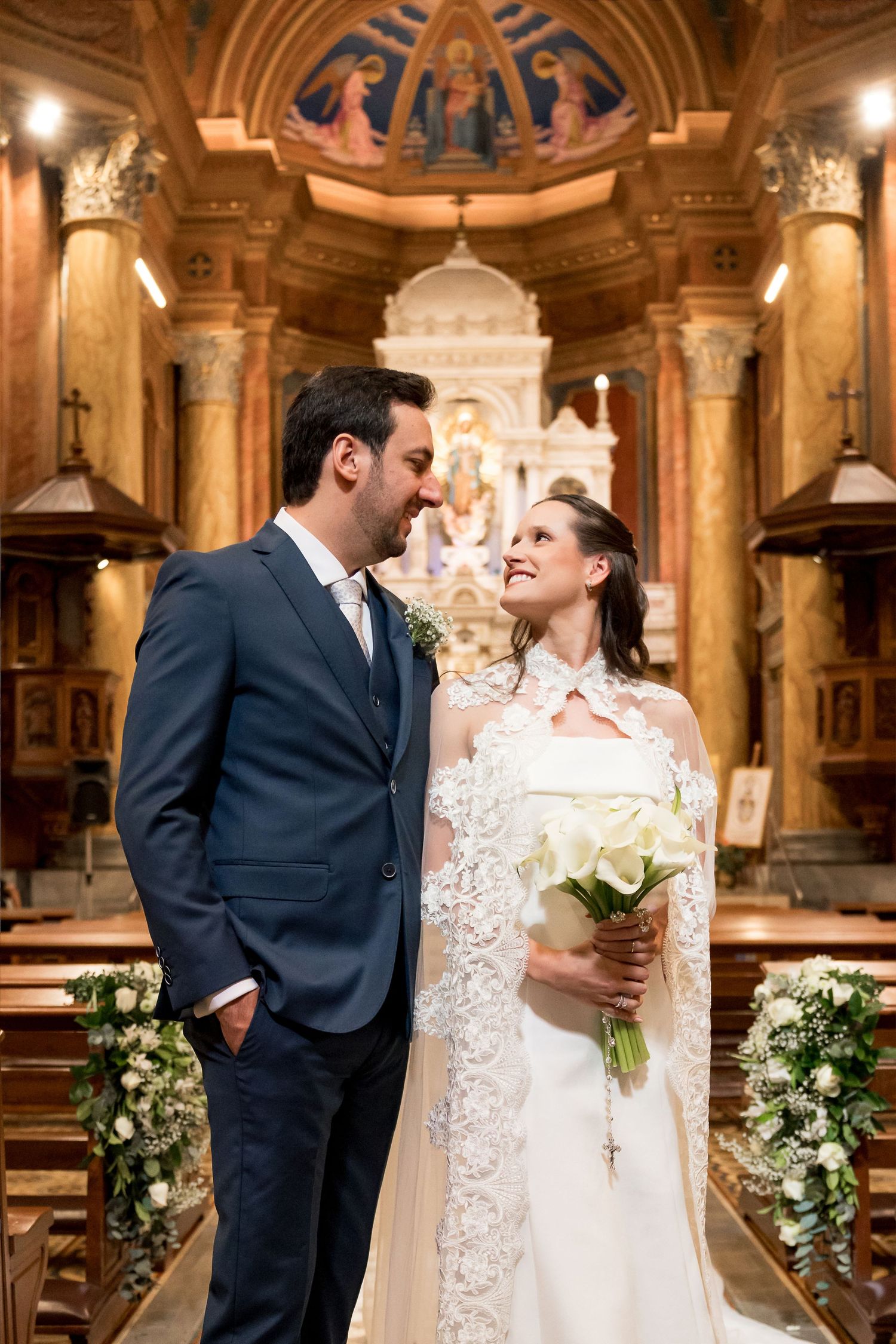 Bride and groom share a loving glance inside an ornate church, bride holding white calla lily bouquet wearing lace cape.