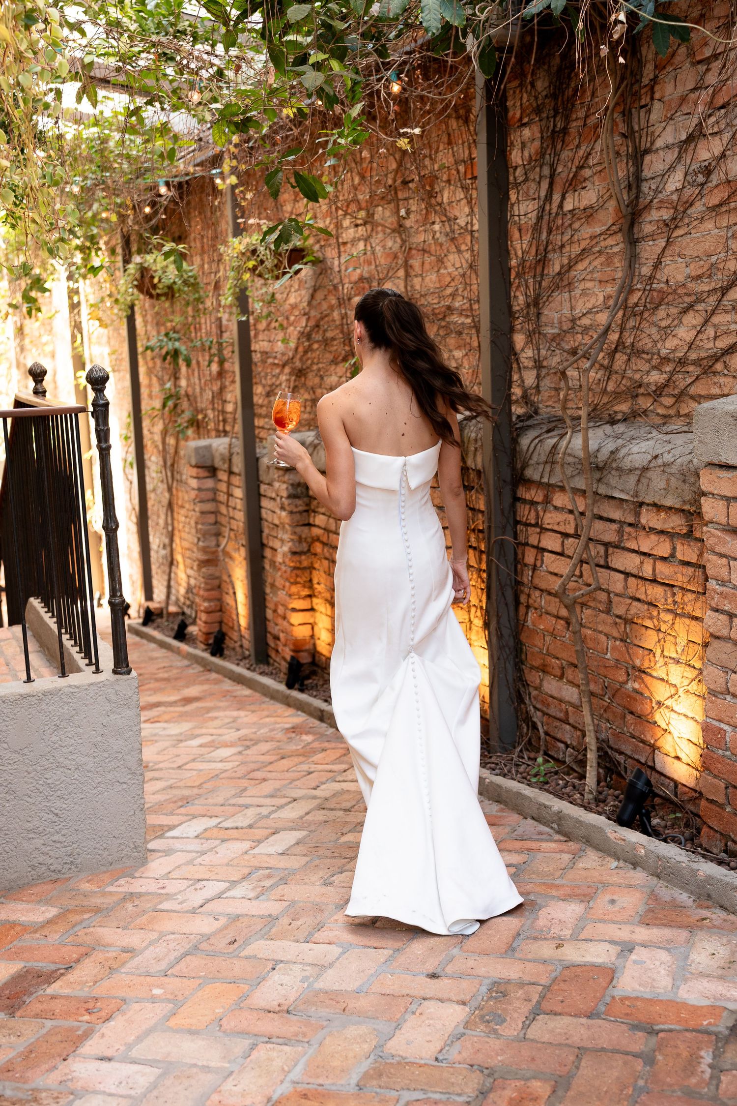 Bride in white strapless gown holding a drink, standing on brick pathway beside illuminated stone wall.