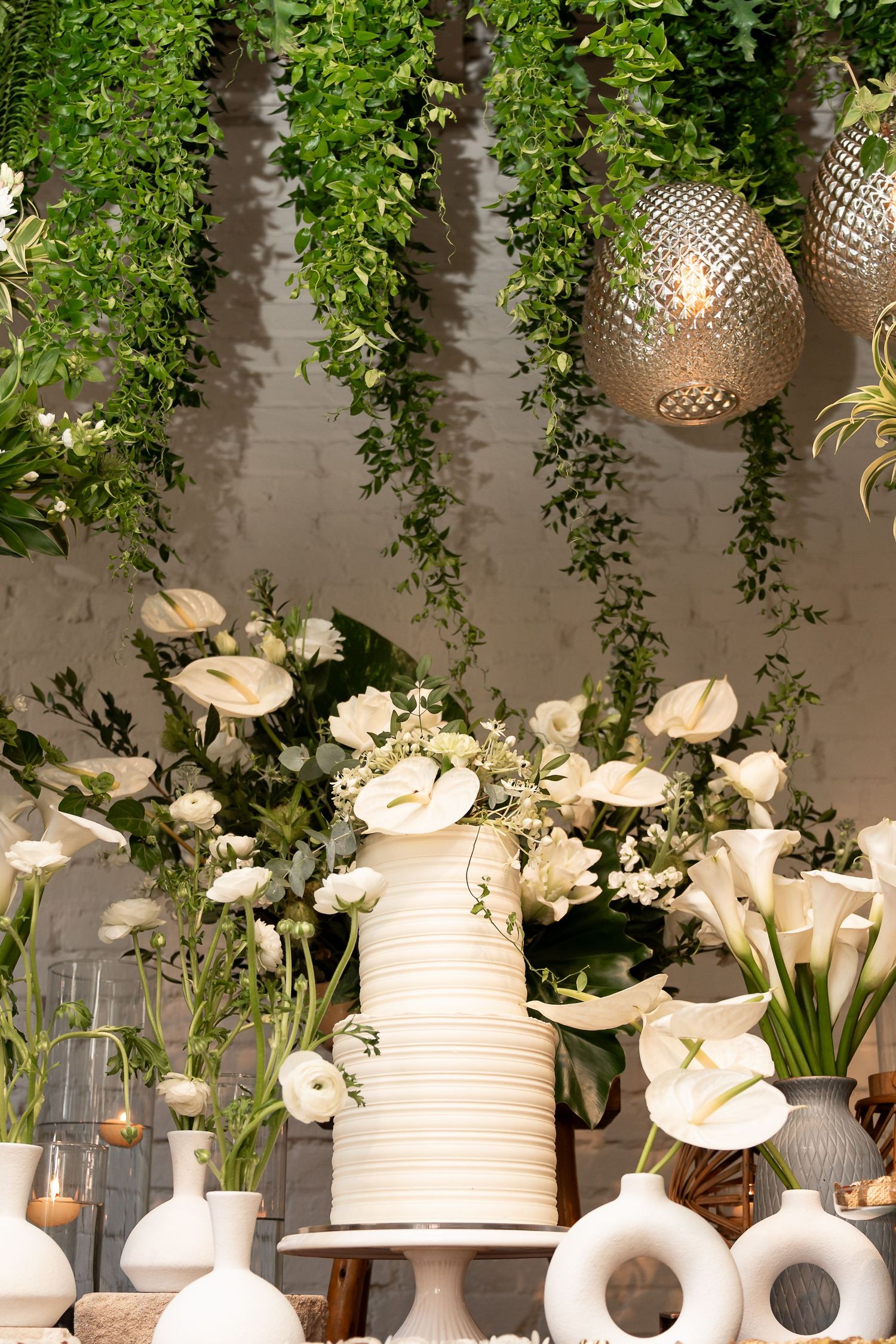 Elegant white wedding cake with calla lilies, surrounded by floral arrangements and hanging greenery in a sophisticated setting.