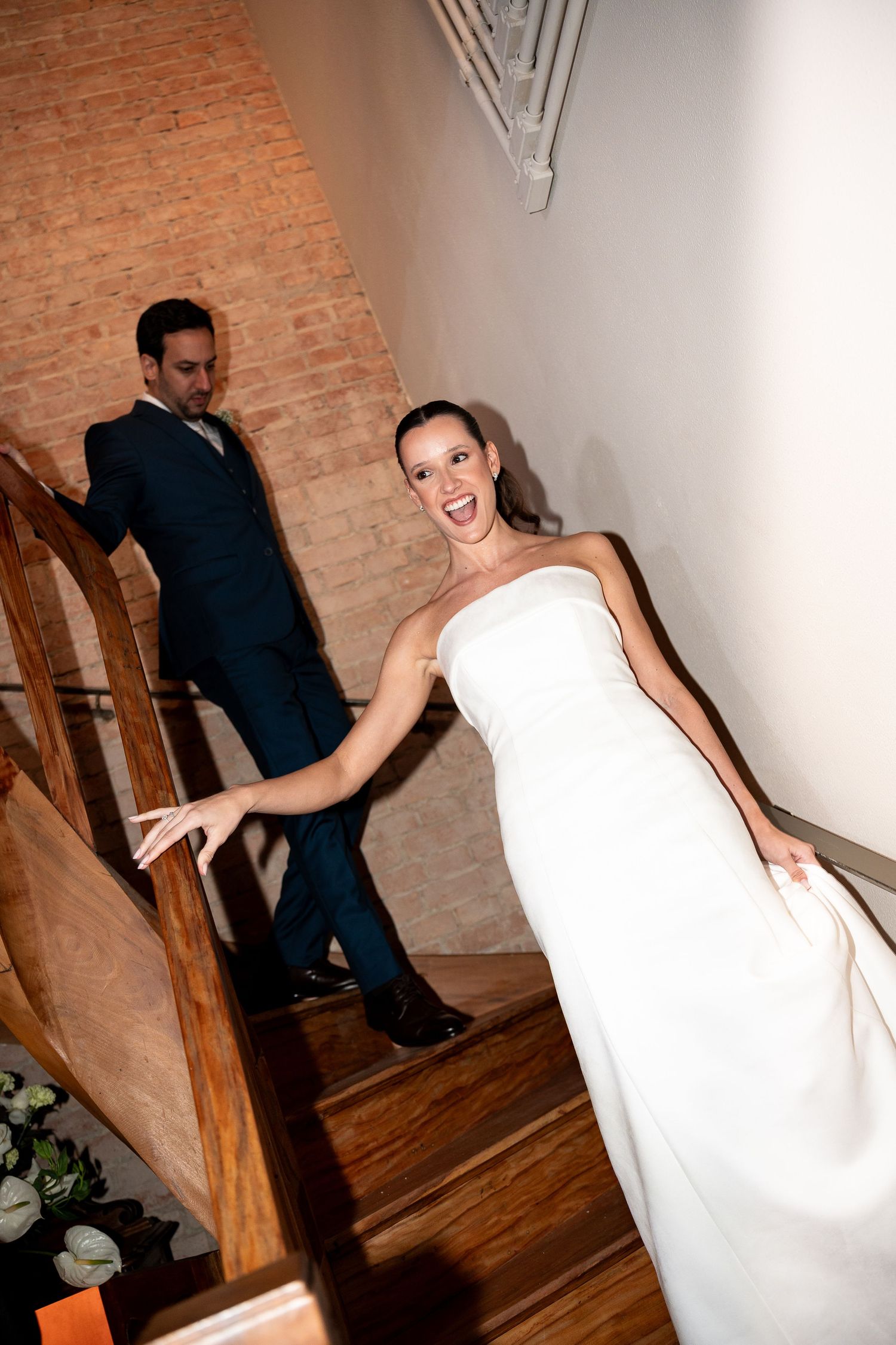 Bride in white strapless gown laughing on wooden staircase with groom in navy suit behind her.