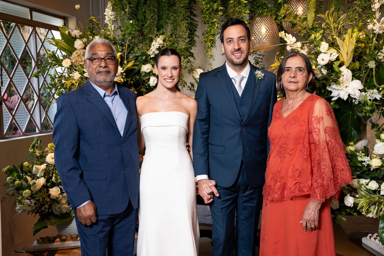 Wedding couple in white dress and blue suit posing with family members before floral backdrop.