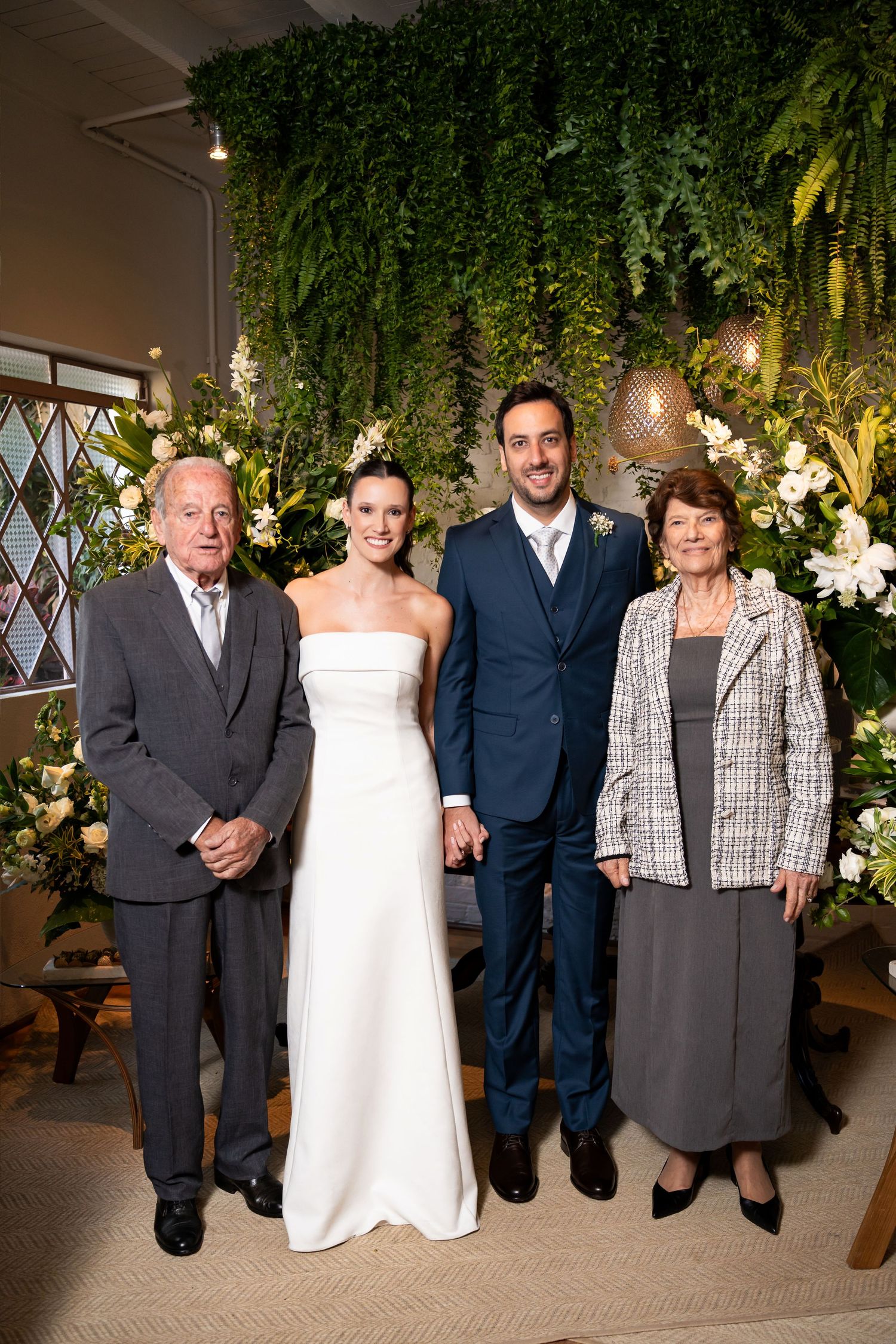 Wedding couple poses with elderly family members in front of lush floral backdrop with white flowers and greenery.