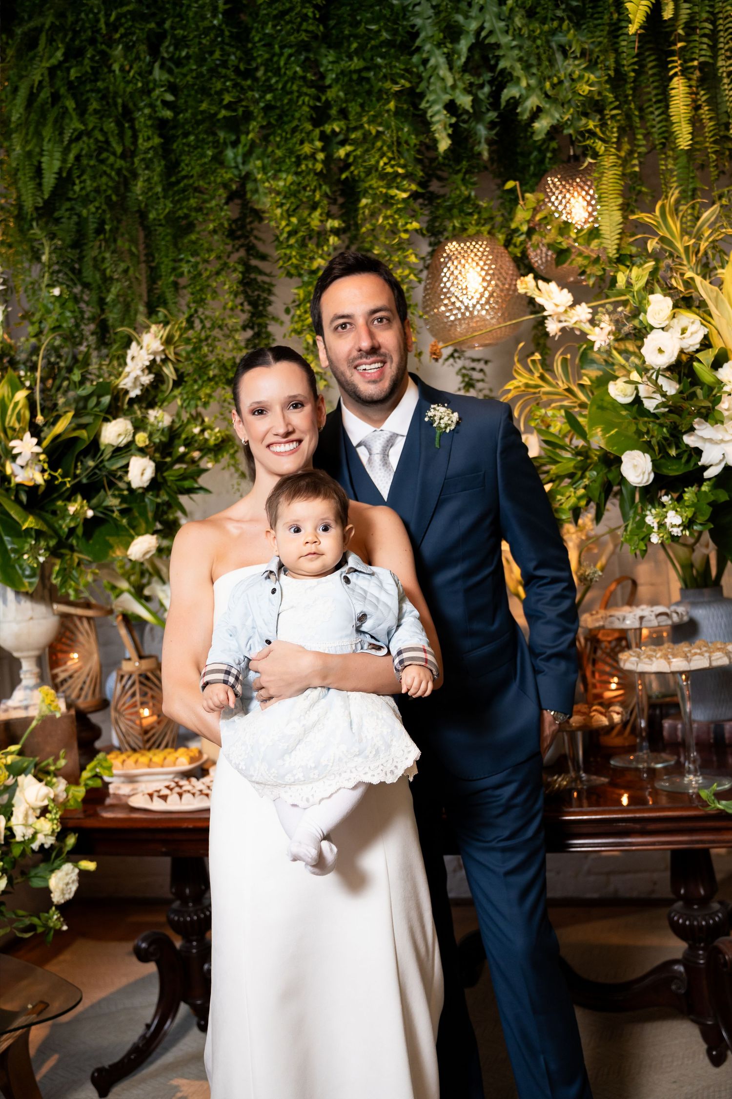 Happy couple in wedding attire holding their baby boy, posing in front of lush green floral backdrop.