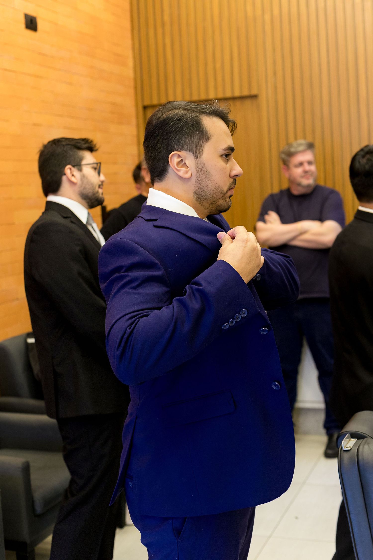 Man in blue double-breasted suit adjusting jacket in conference room with colleagues in background.