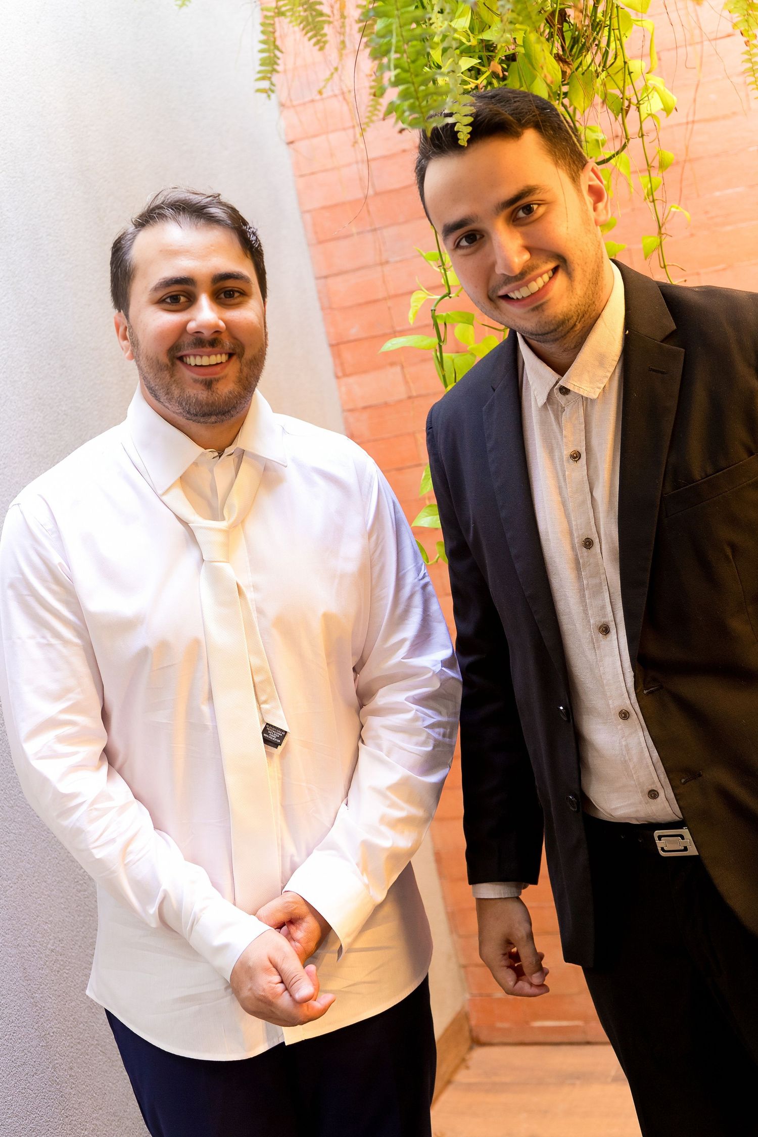 Two smiling men in formal attire pose together indoors near a brick wall with green plants.