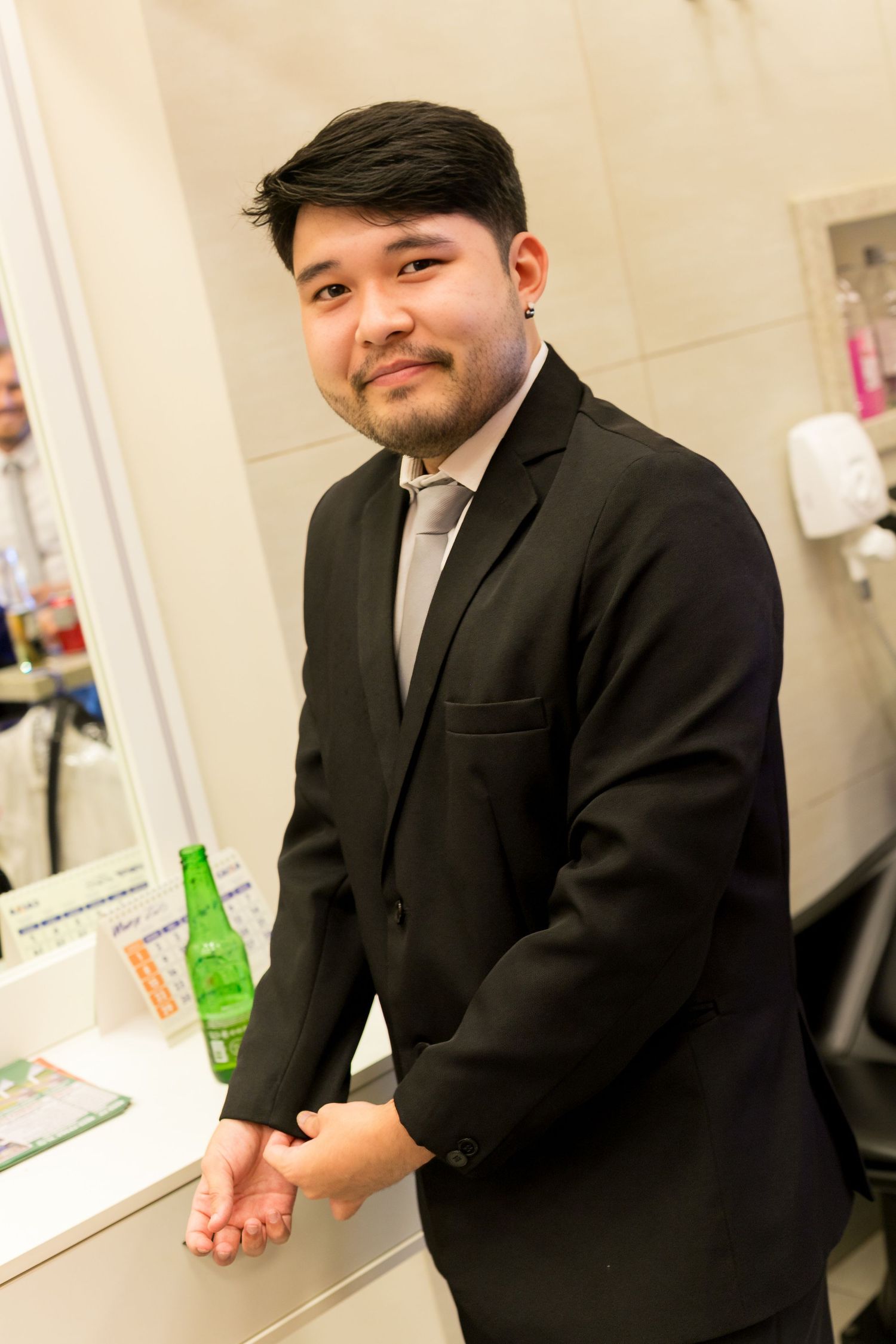 Young man in black suit and gray tie smiling in a bathroom setting with a green bottle nearby.
