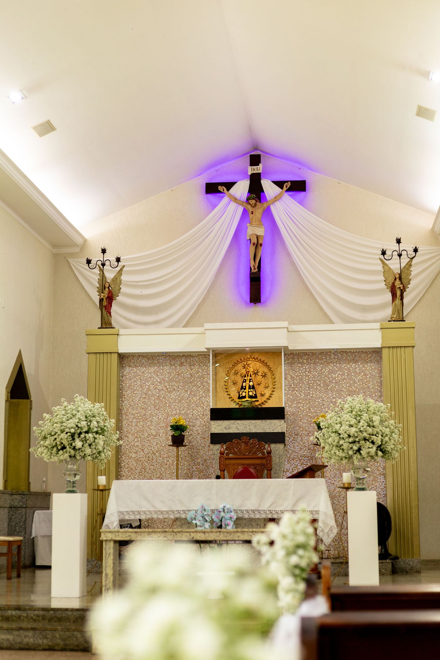Church altar with crucifix illuminated by purple light, white draping, floral arrangements, and religious statues on a decorated sanctuary.