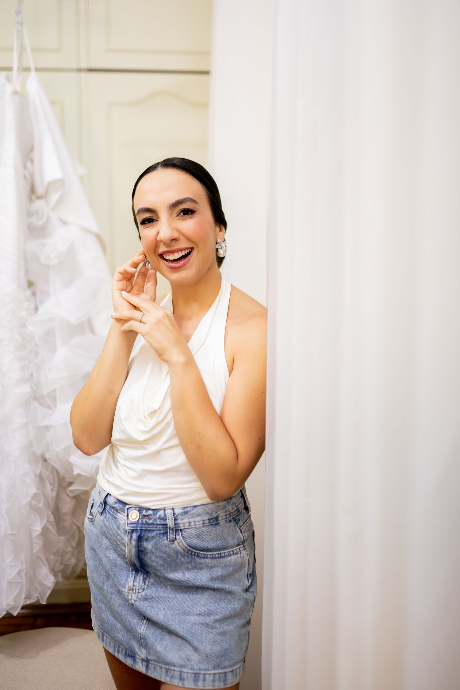 Smiling bride-to-be trying on earrings in a bridal boutique, wearing a white top and denim skirt.