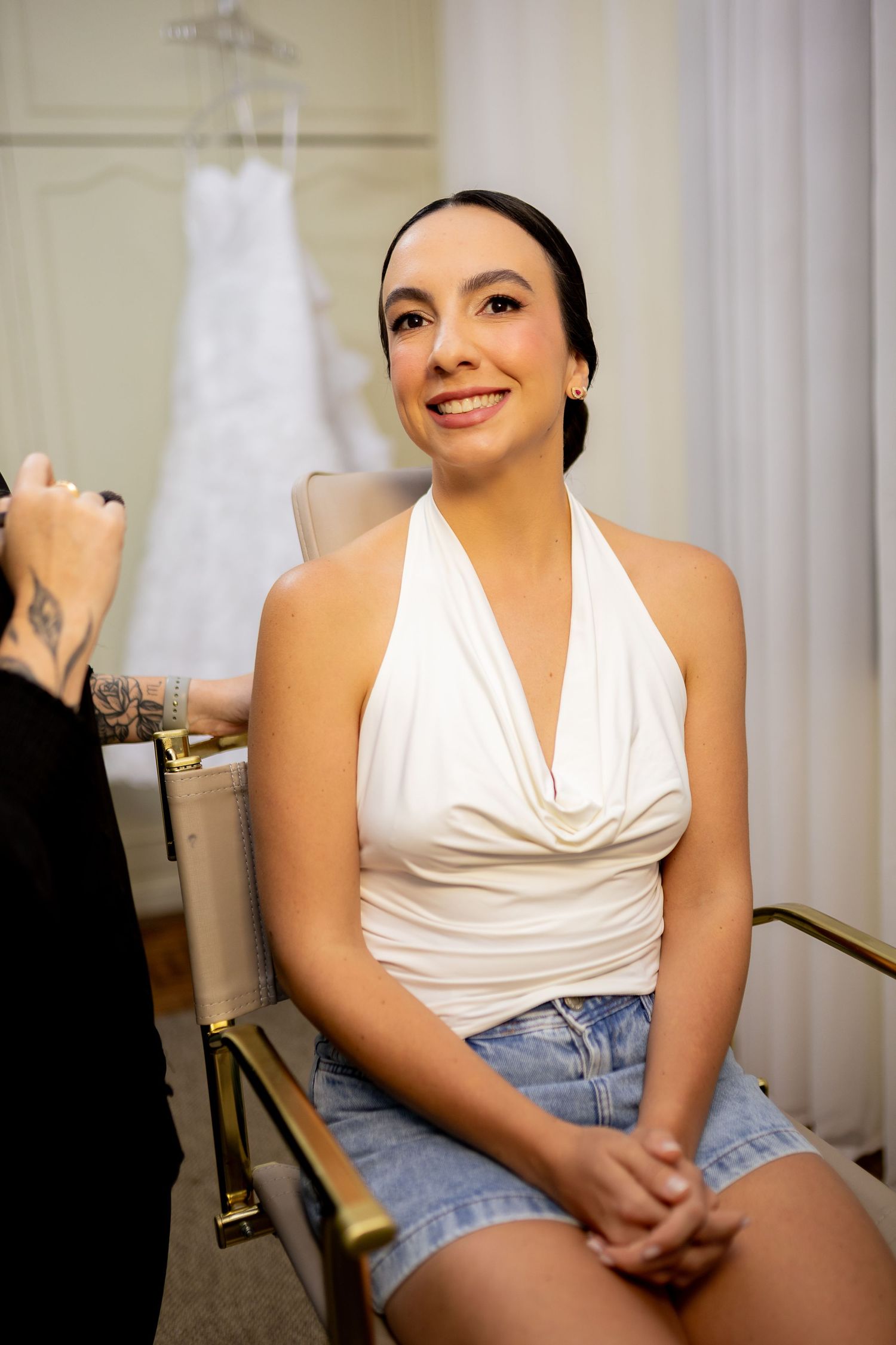 Smiling bride-to-be in white halter top and denim skirt sits in chair during bridal prep, wedding dress visible in background.