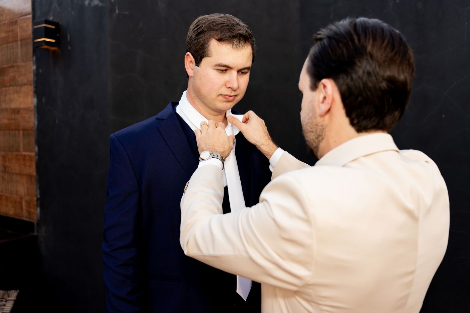 A man in a navy suit having his white tie adjusted by another man in a cream shirt.
