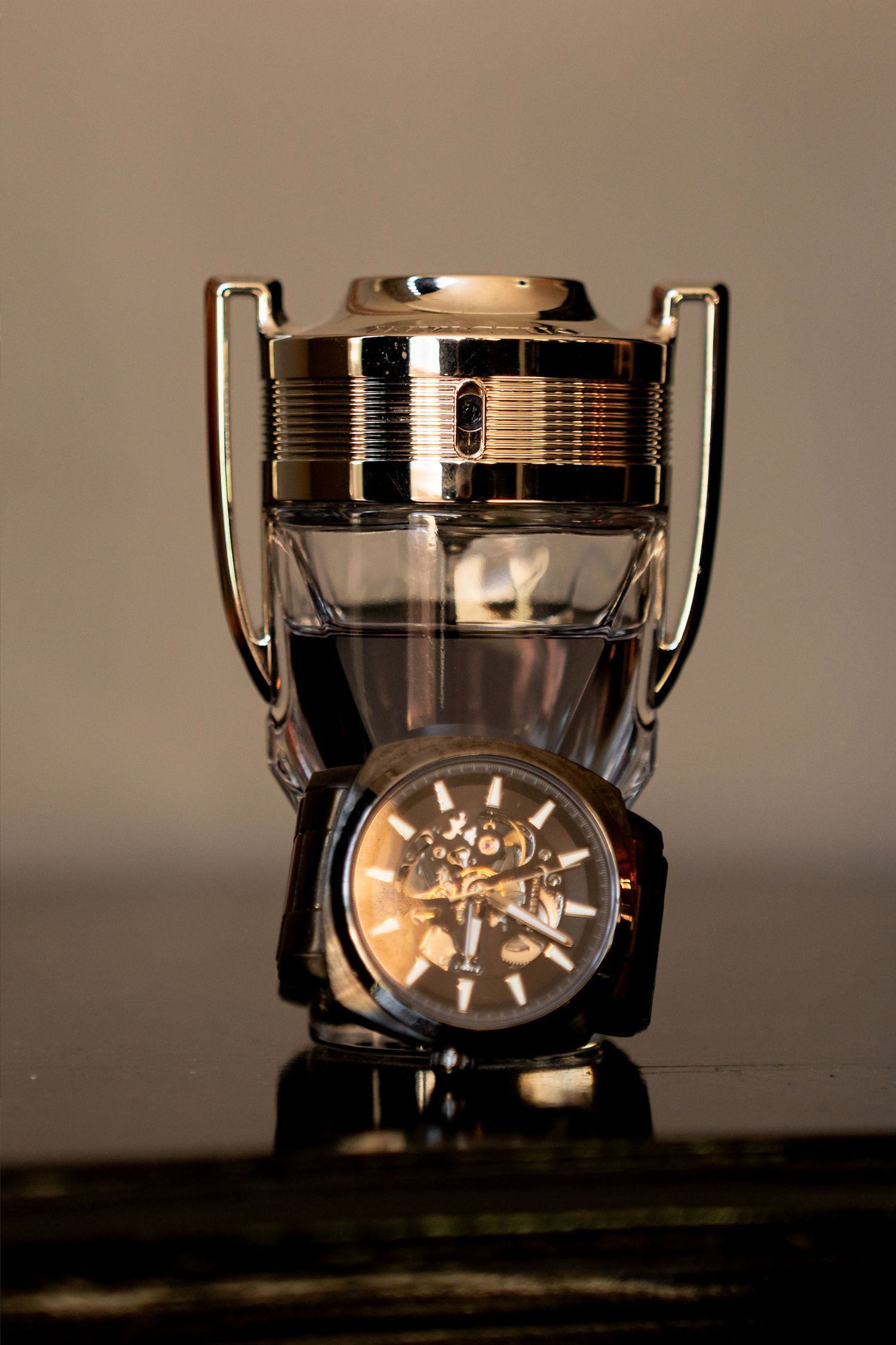 A luxury skeleton watch displayed in front of a trophy-shaped glass perfume bottle on a reflective surface.
