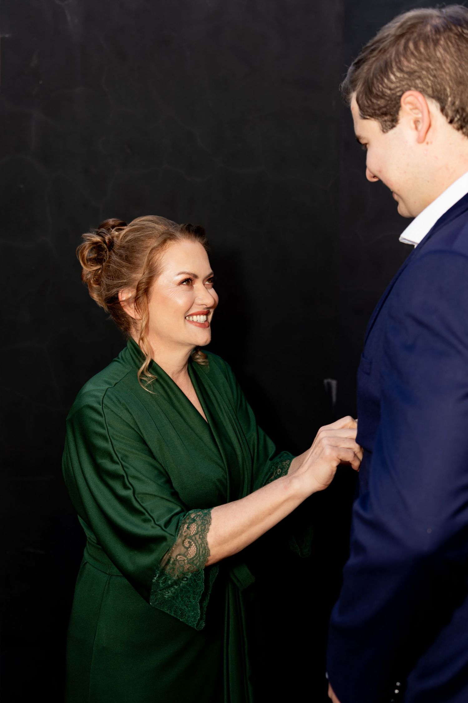 A smiling woman in a green satin robe adjusts a man's navy suit jacket against a dark background.
